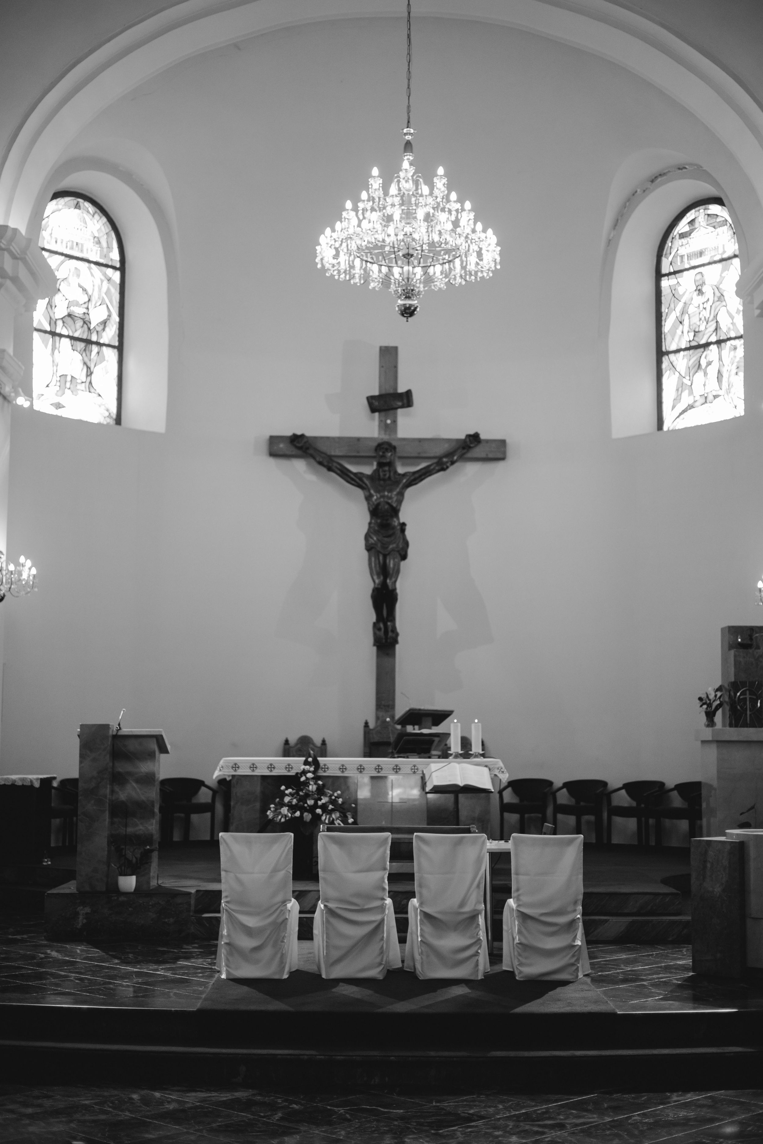 Black and white photo of a church altar with crystal chandelier and crucifix beneath arched windows.