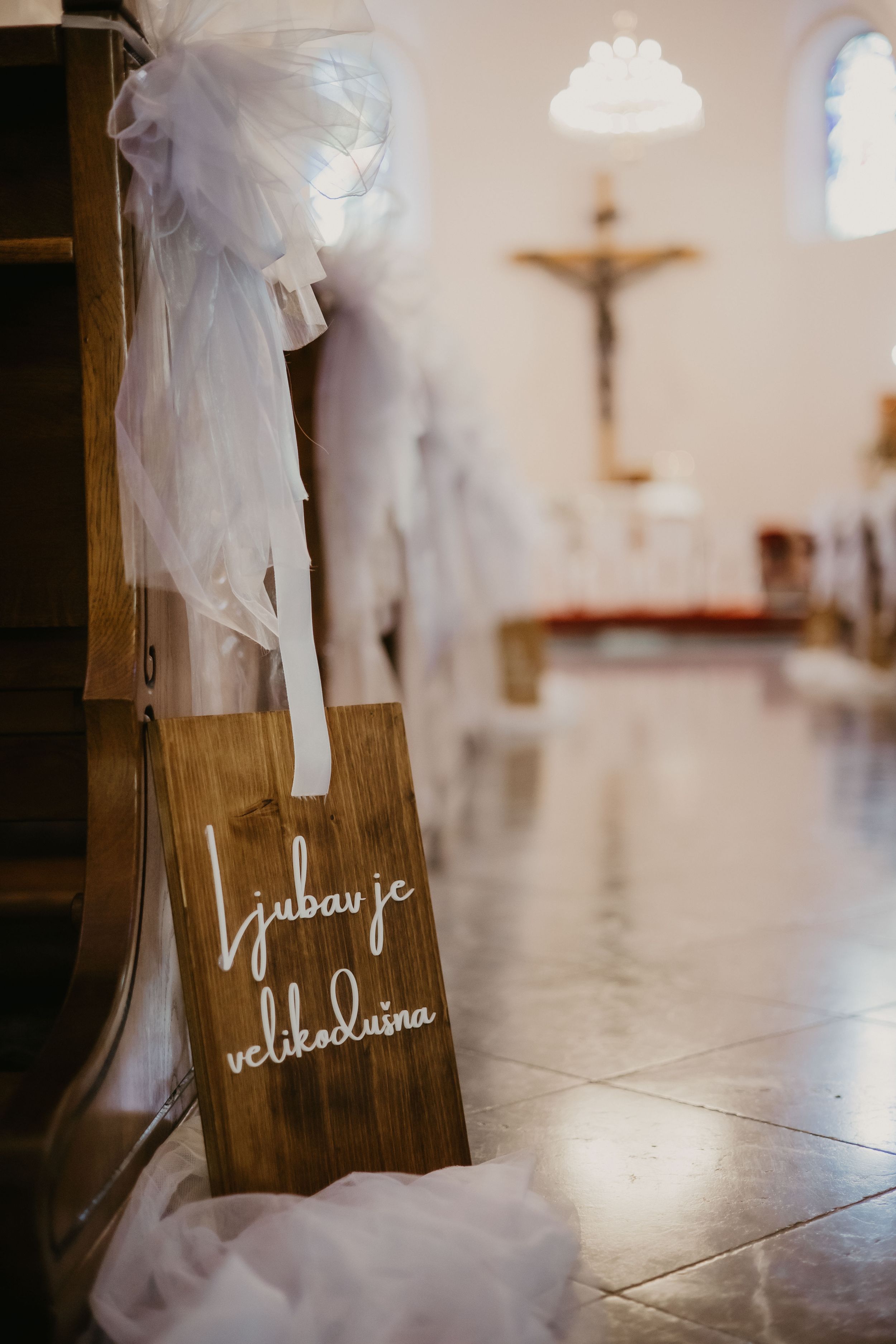 A wooden sign with text stands in a church interior with tulle fabric draping and a crucifix in the background.