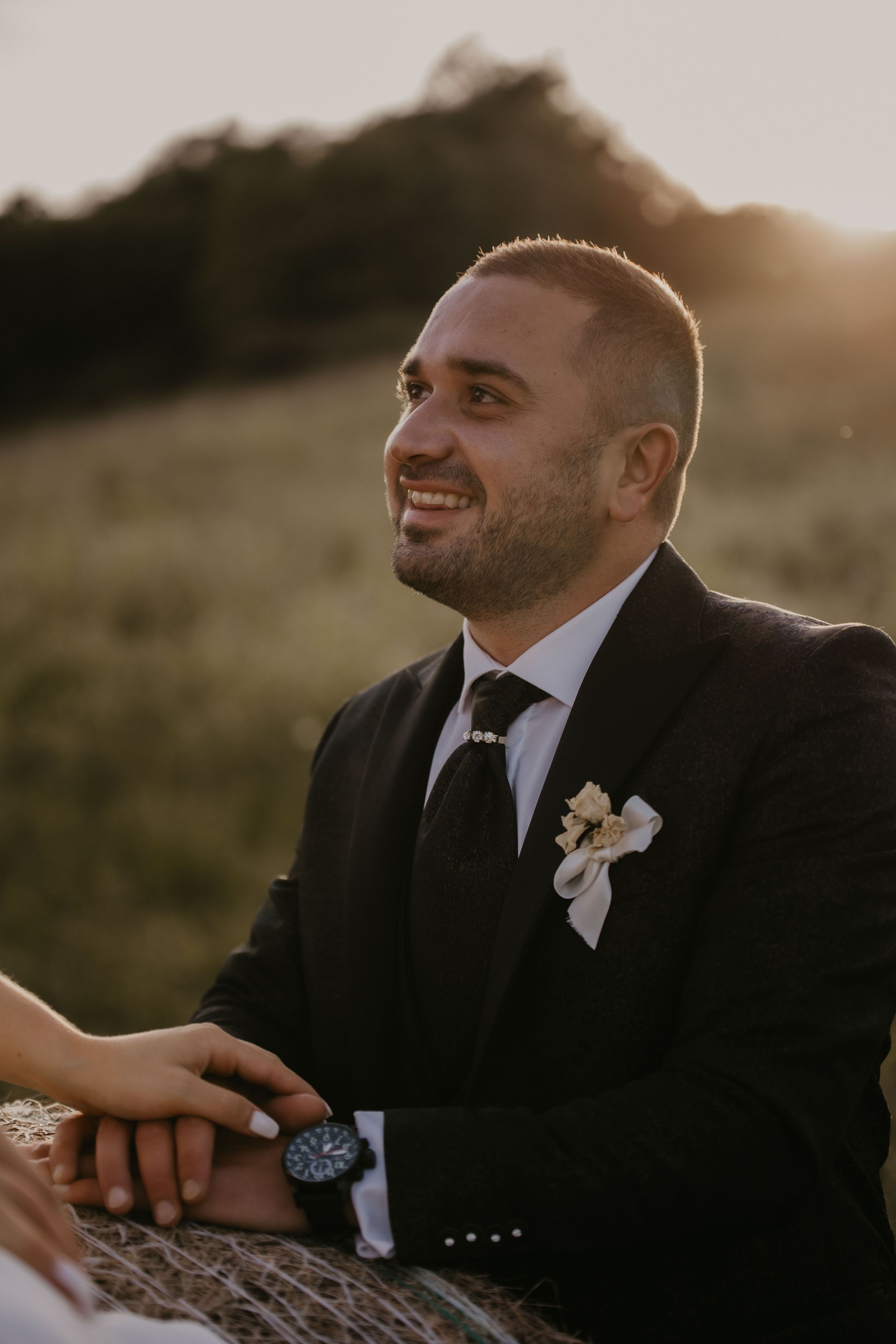 A groom wearing a black suit and boutonniere smiles warmly during an outdoor sunset wedding photo session.