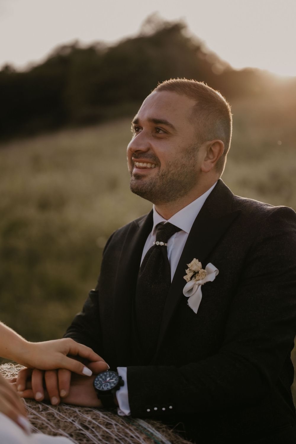 A groom wearing a black suit and boutonniere smiles warmly during an outdoor sunset wedding photo session.