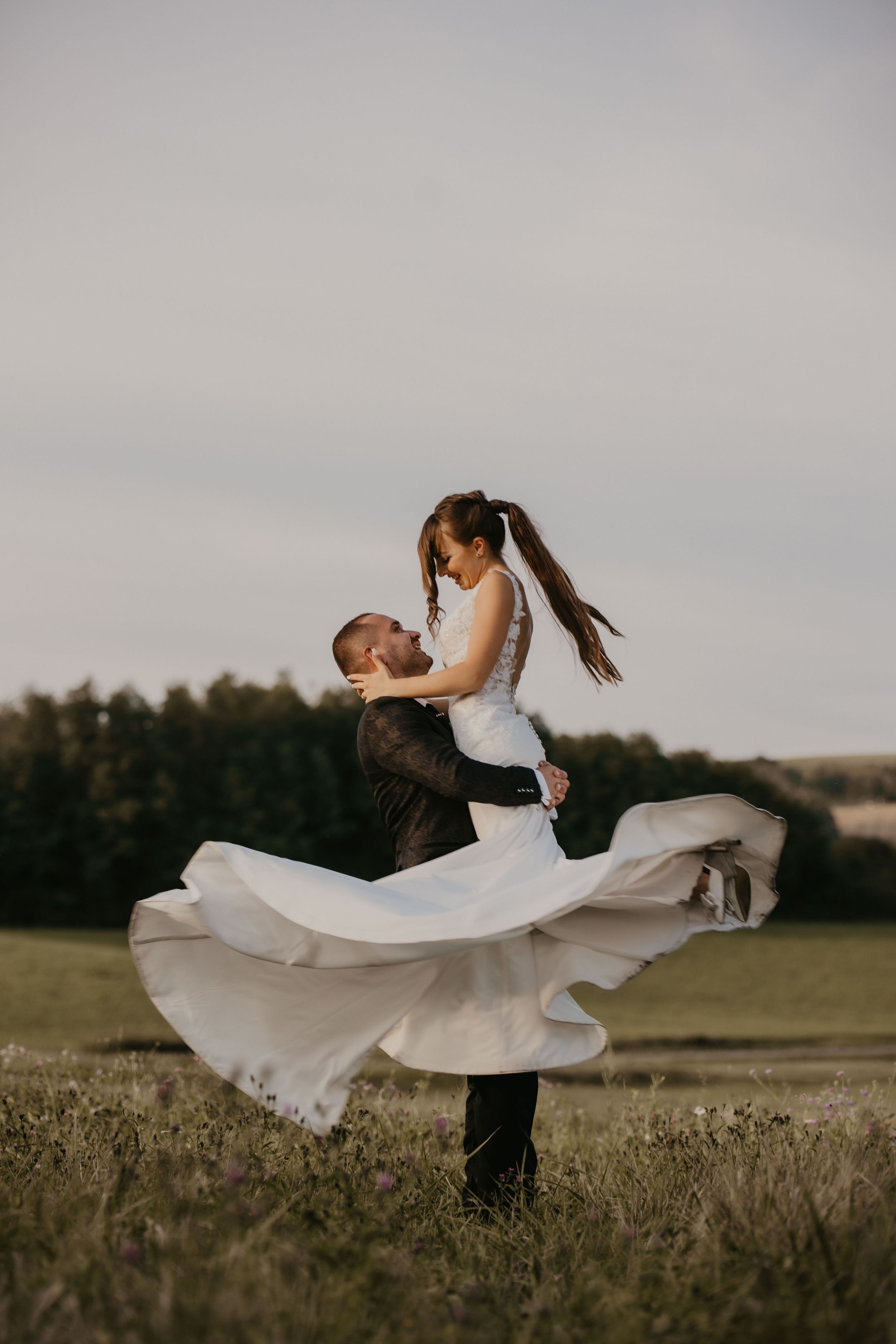 Groom lifts bride in flowing white dress as her dress twirls dramatically in an outdoor field at sunset.