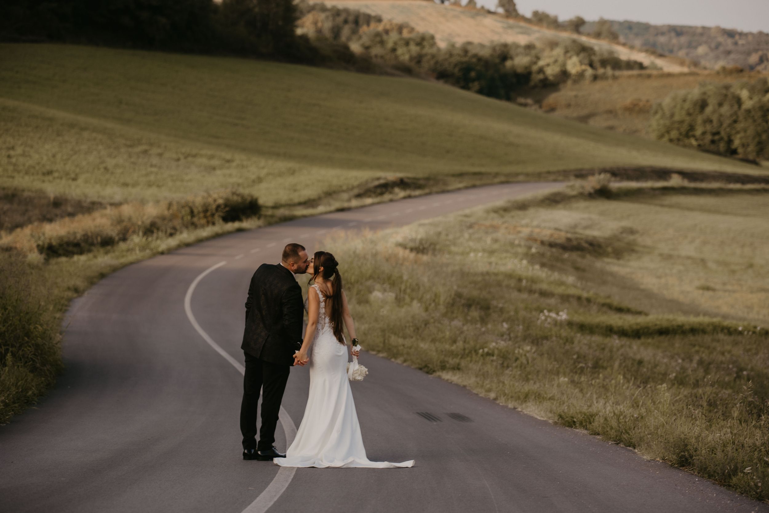 Newlyweds stand together on a winding country road surrounded by rolling hills.