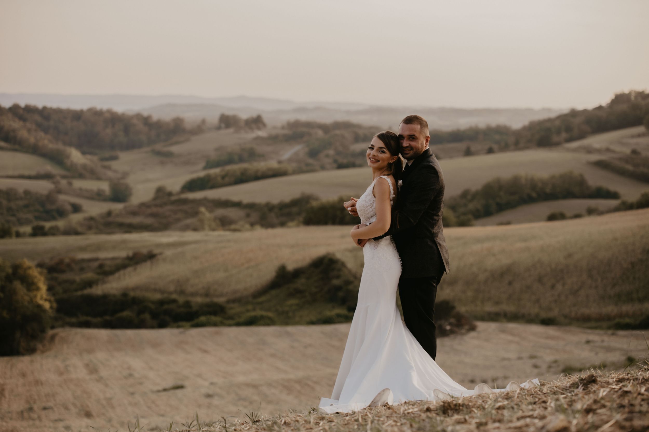 Wedding couple embraces while standing on hilltop overlooking scenic valley at sunset.