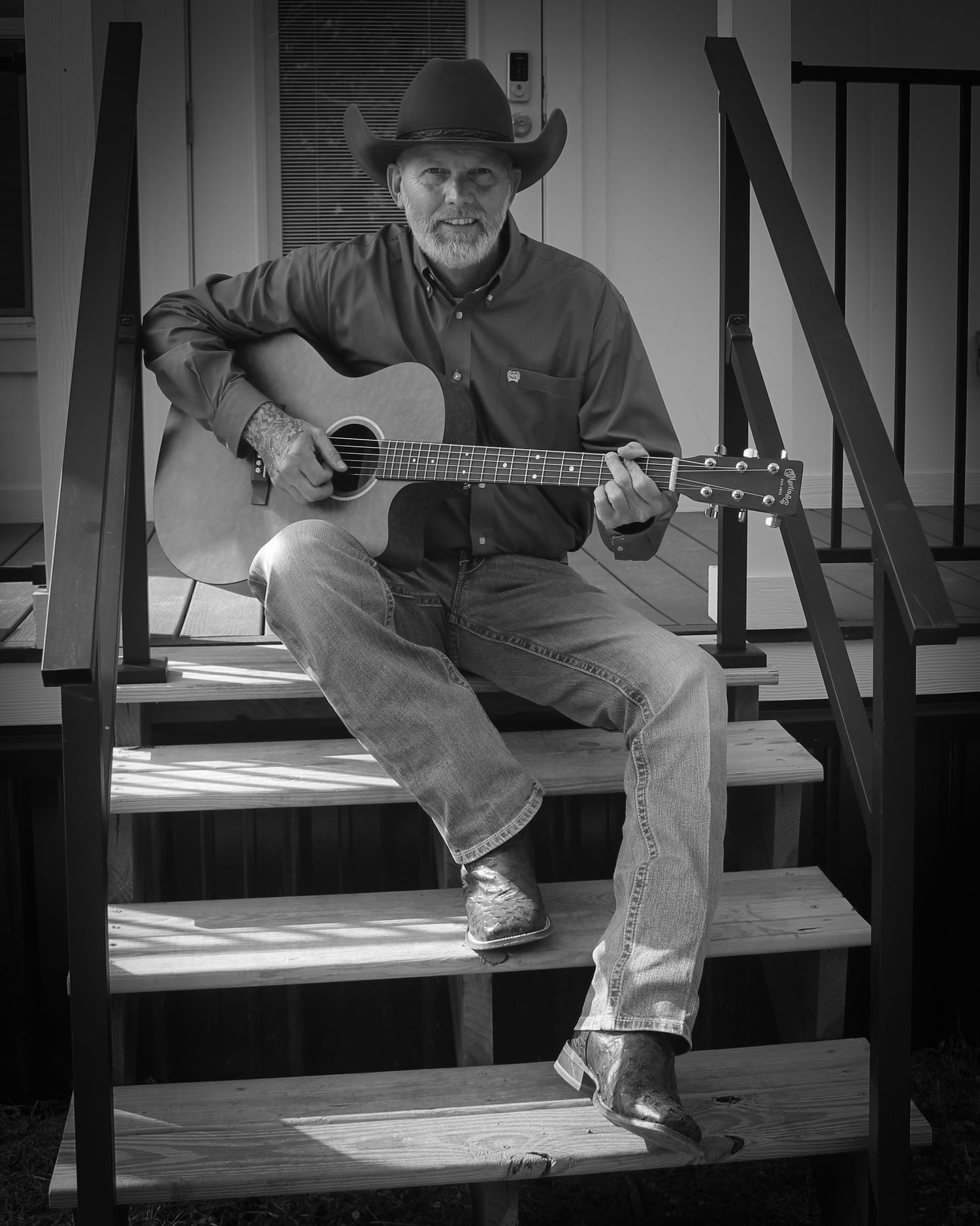 Black and white portrait of musician in cowboy hat playing acoustic guitar while seated on porch steps.