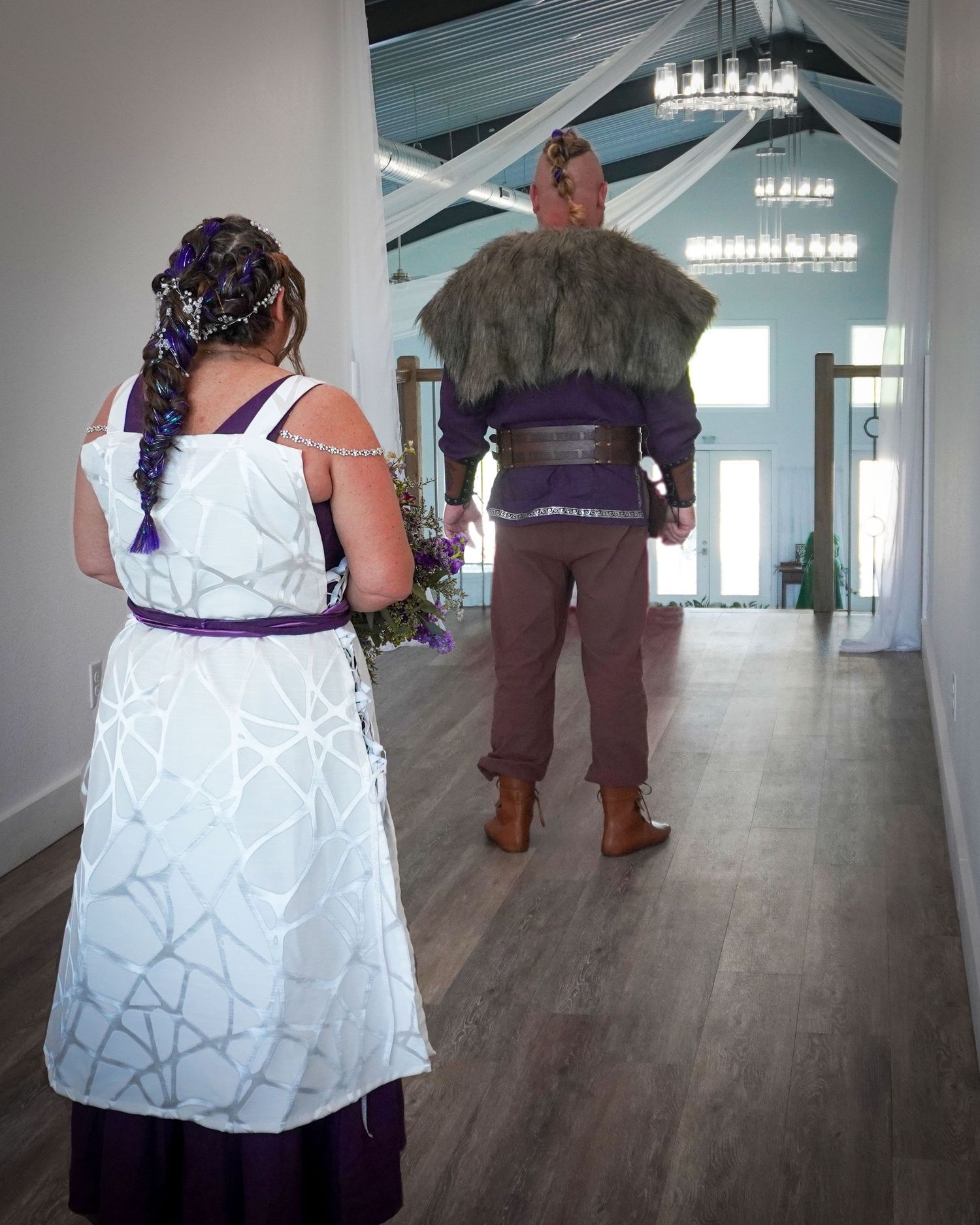 A couple in coordinated purple and white wedding attire stand in an elegant venue hallway.