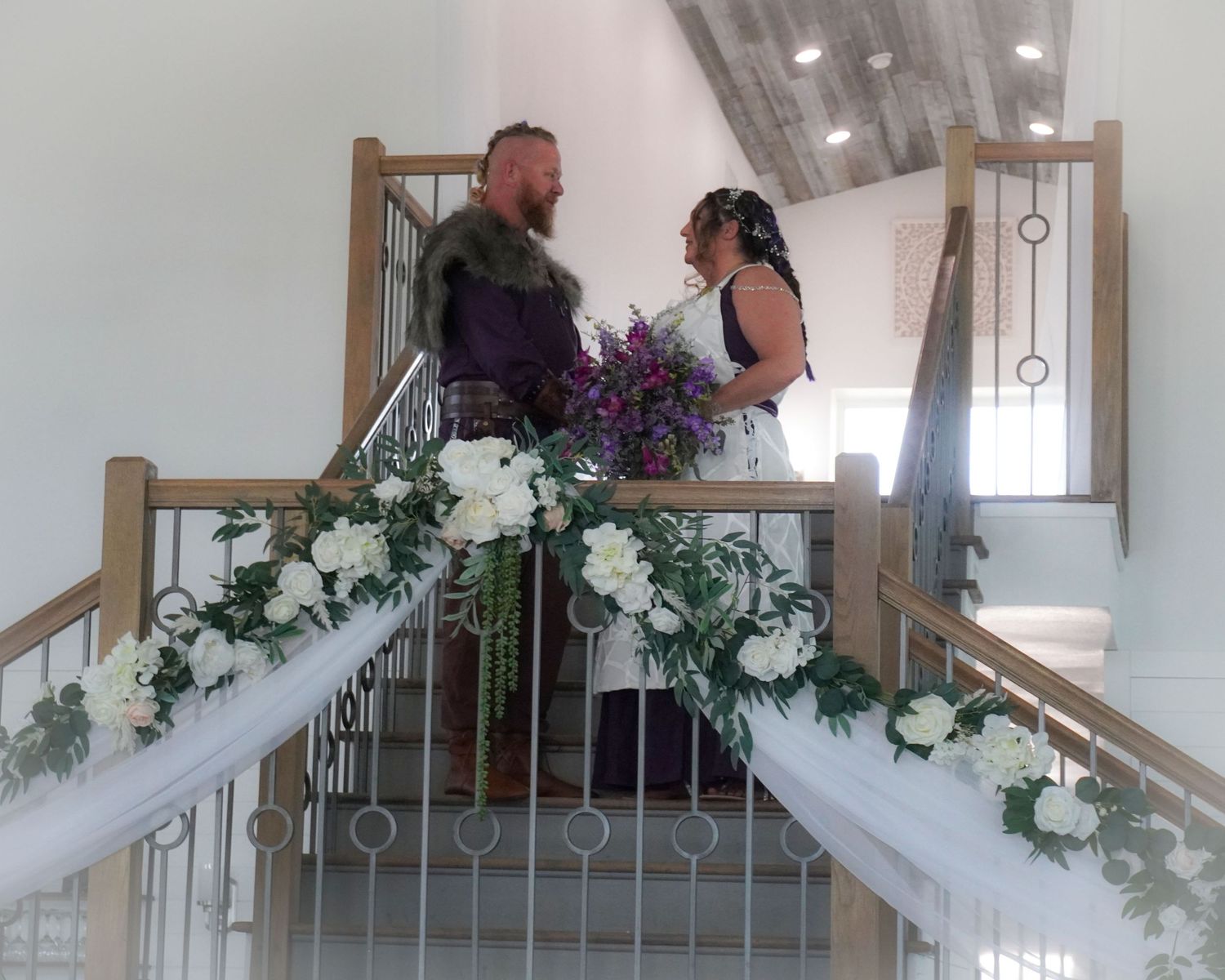 Wedding couple poses on elegant staircase decorated with white and purple floral arrangements and draped fabric.