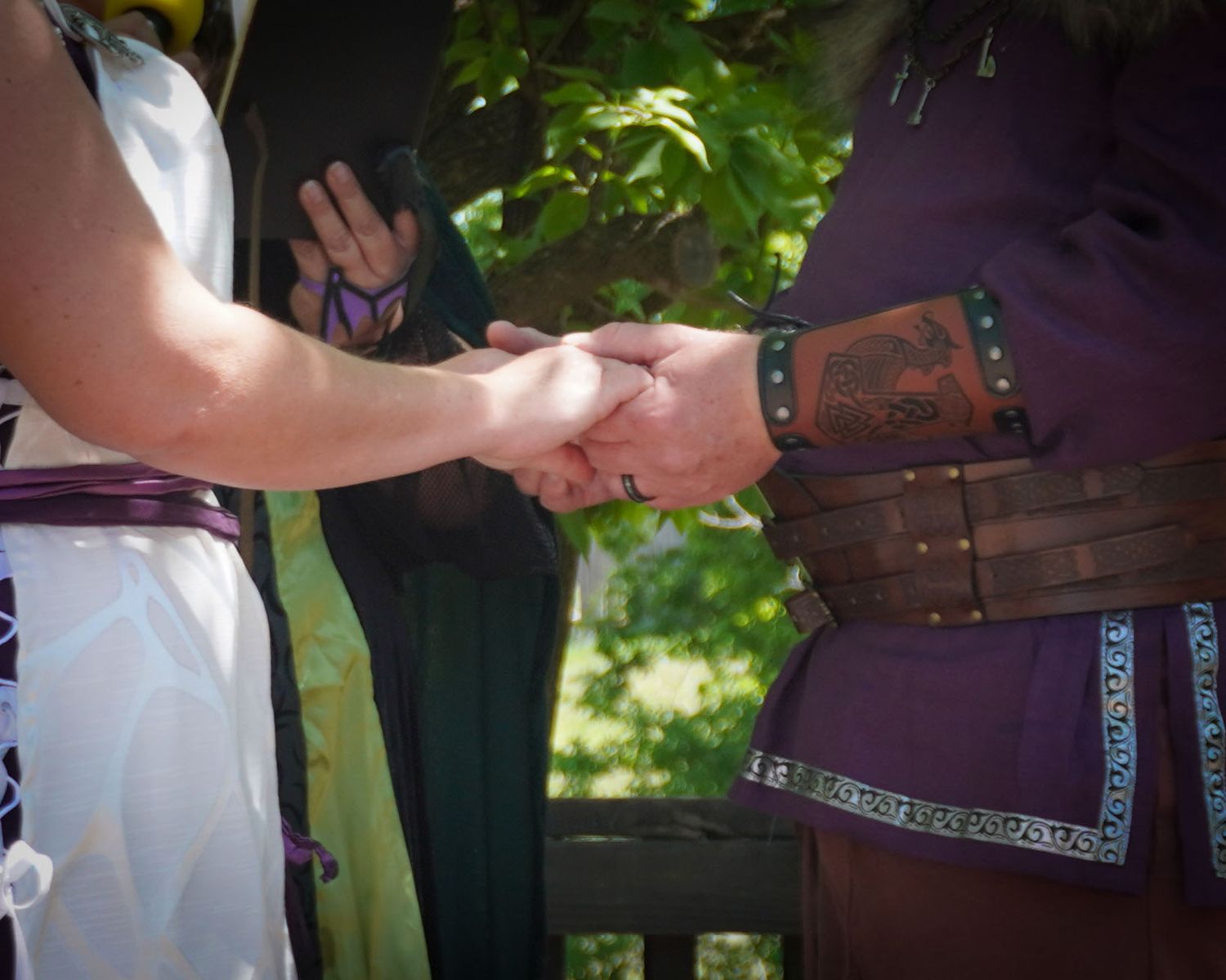 Two people wearing medieval costumes shake hands outdoors with sunlit trees in the background.