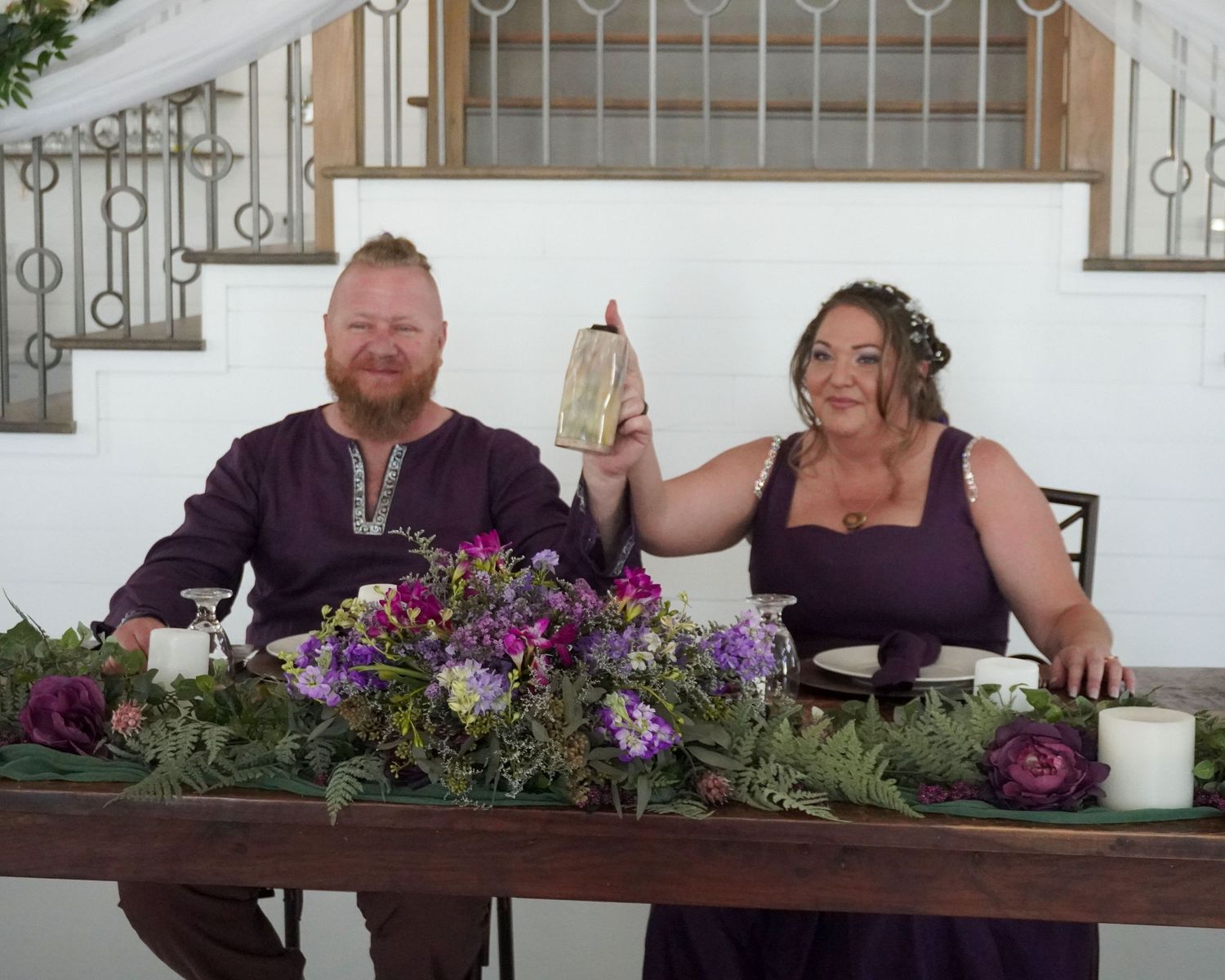 Wedding couple toasts at a reception table decorated with purple flowers and rustic decor.