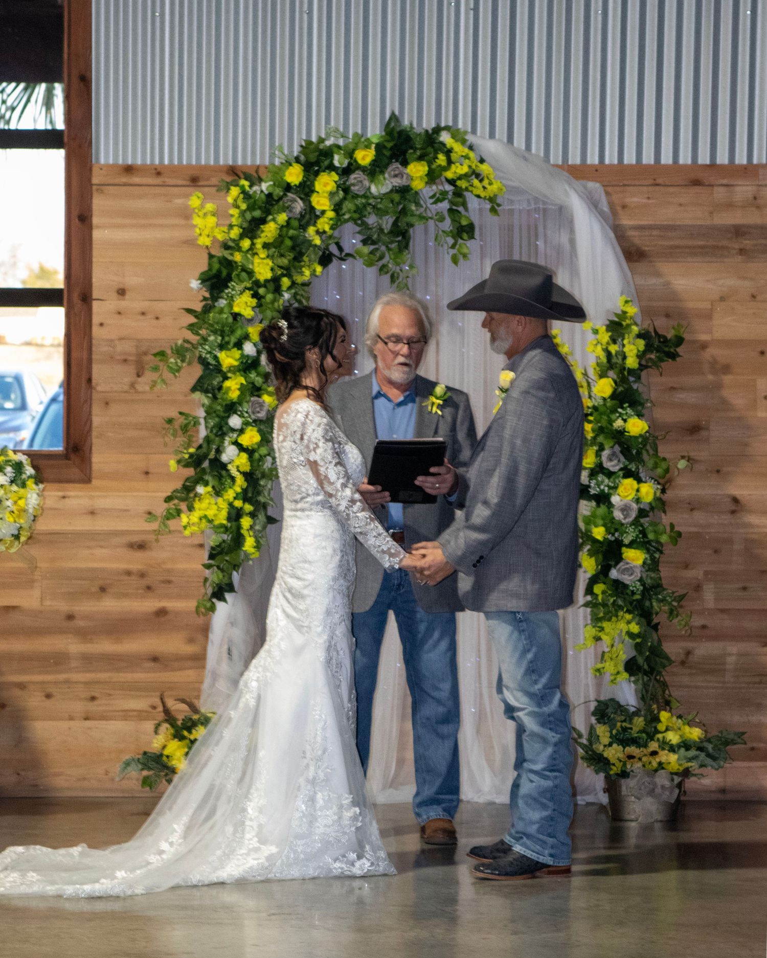 A wedding ceremony takes place under a beautiful floral arch with yellow blooms against a wooden wall backdrop.