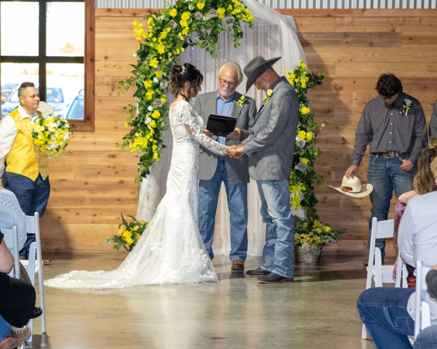 Rustic country wedding ceremony taking place in front of a wooden wall decorated with yellow floral arch.