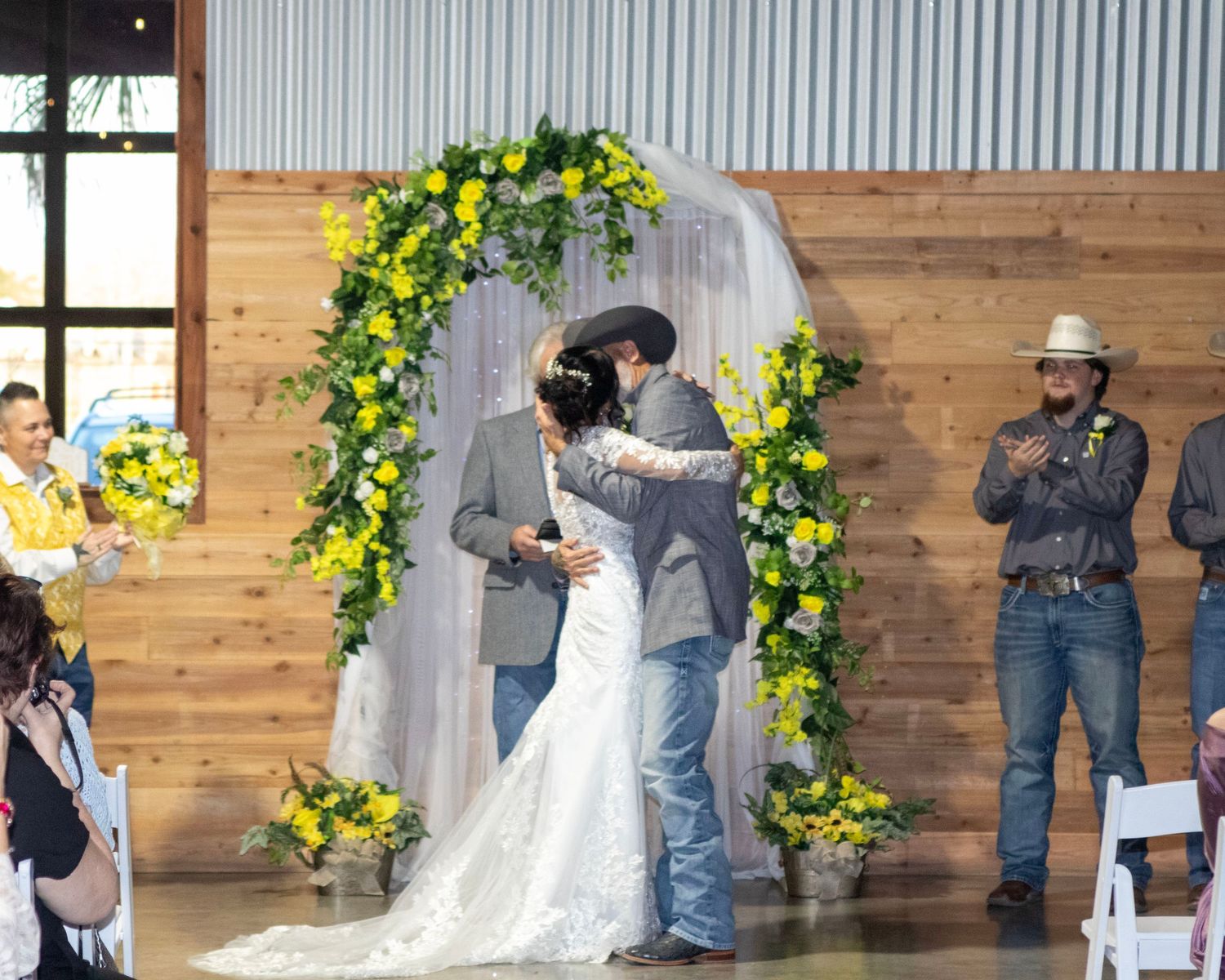Wedding ceremony taking place under a beautiful yellow floral arch in a rustic barn venue.