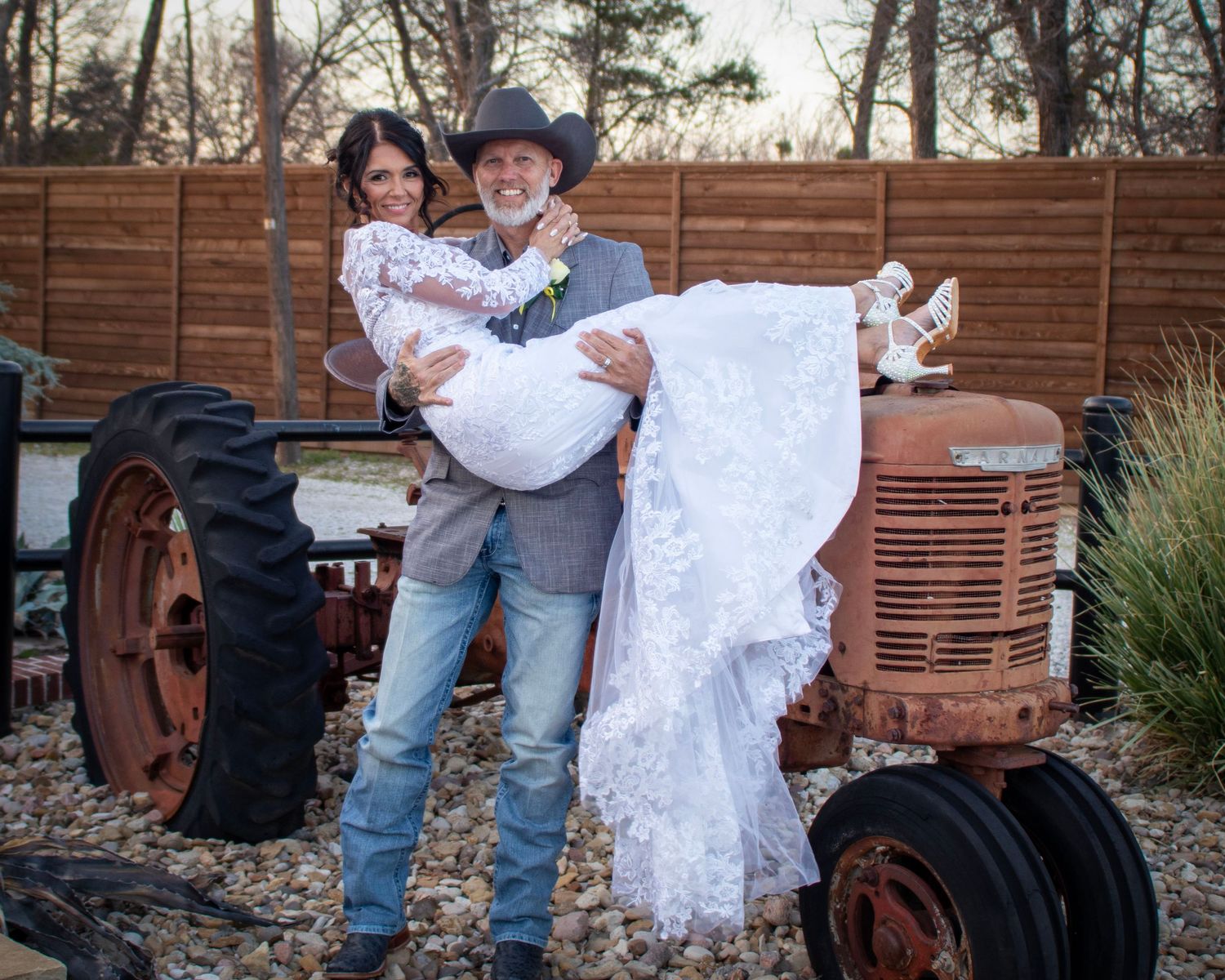 A couple poses playfully in wedding attire in front of a vintage red tractor for a country-themed wedding photo.