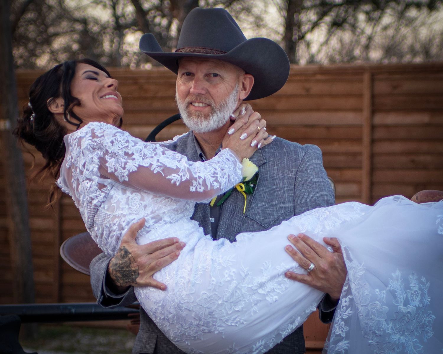 A couple embraces during their wedding ceremony, with the bride wearing a white lace dress and groom in a cowboy hat.