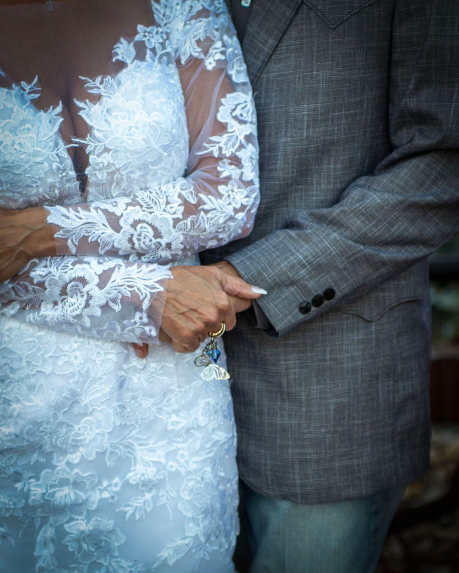 A close-up detail shot of hands and intricate white lace wedding dress next to a grey suit.
