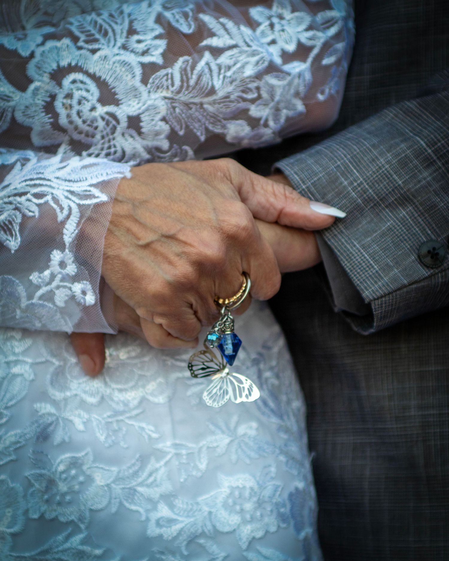 Detailed close-up of hands clasped together showing white lace wedding dress detail and a blue charm accessory.