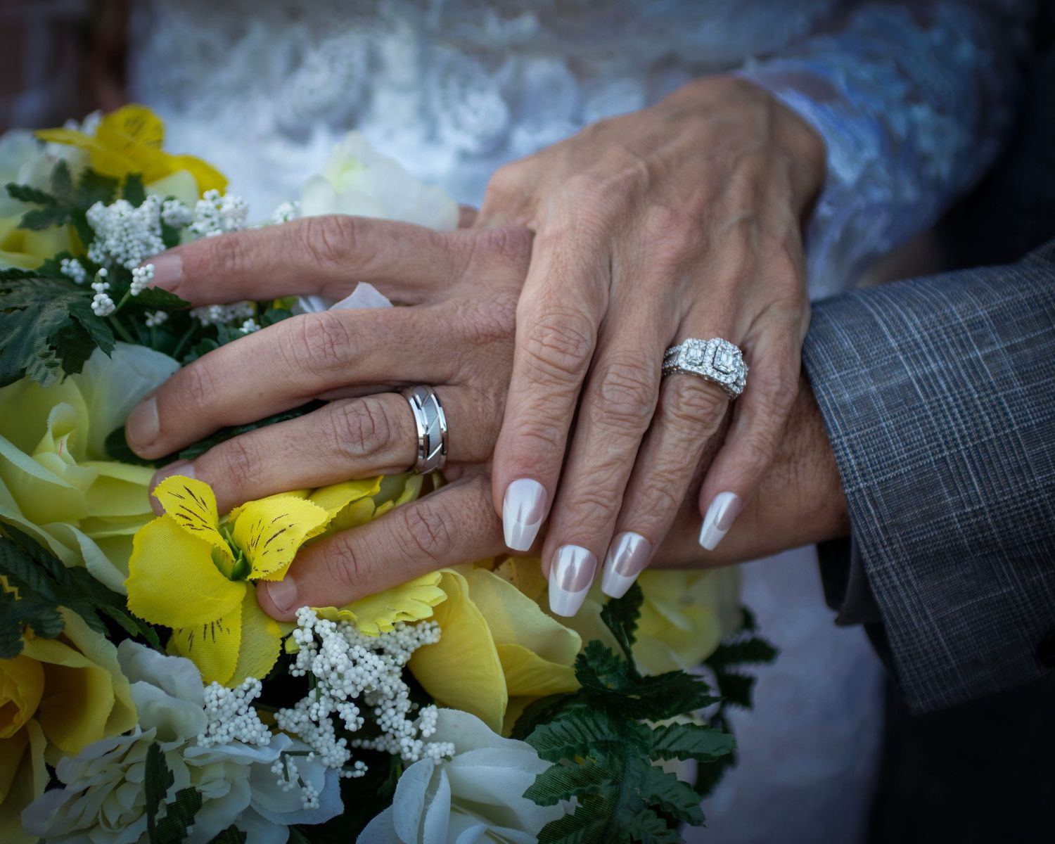 Close-up of newlyweds' hands with wedding rings resting on yellow lily bridal bouquet.