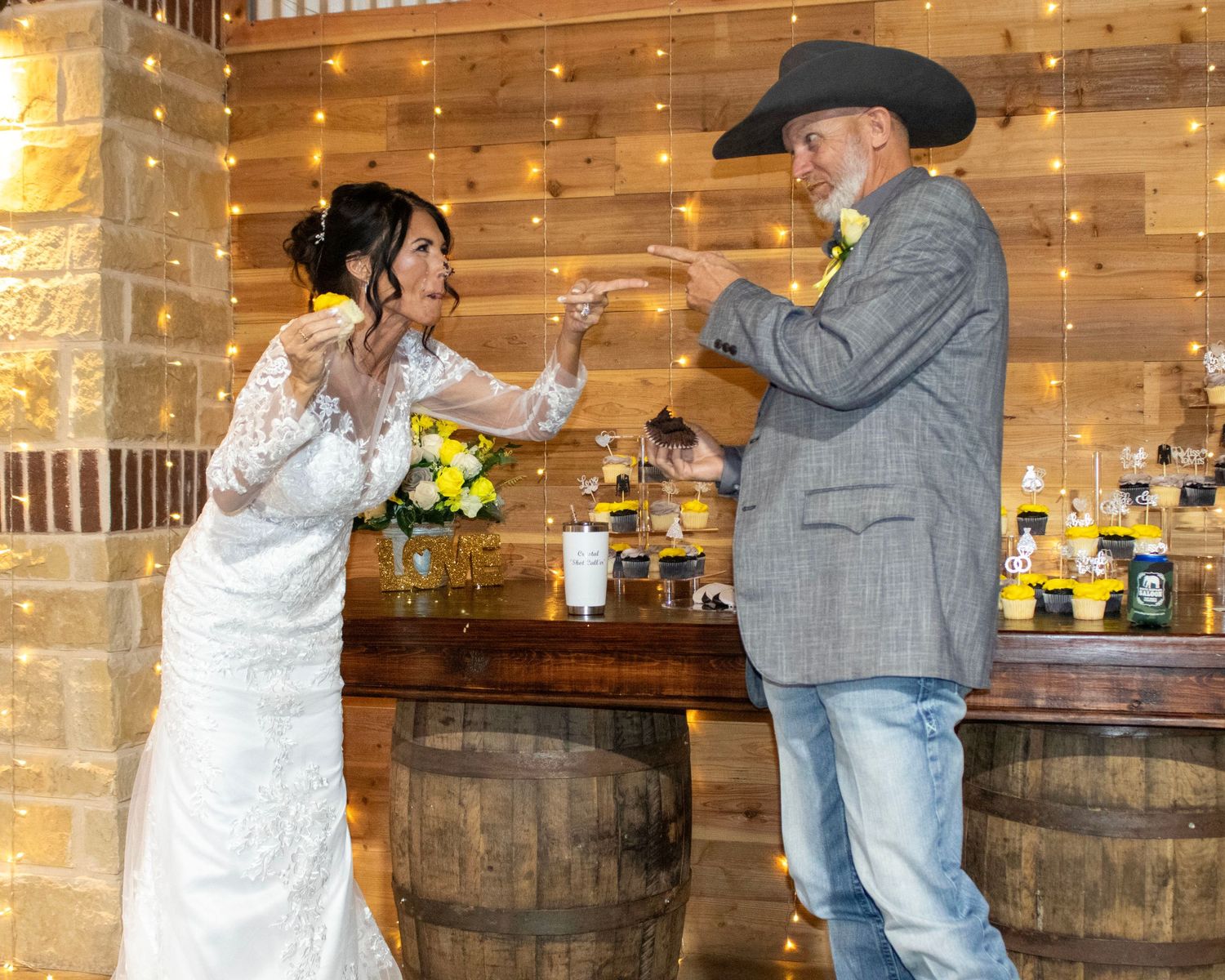 A bride in white lace dress and cowboy share a playful dance moment in a rustic barn setting with wooden barrels.