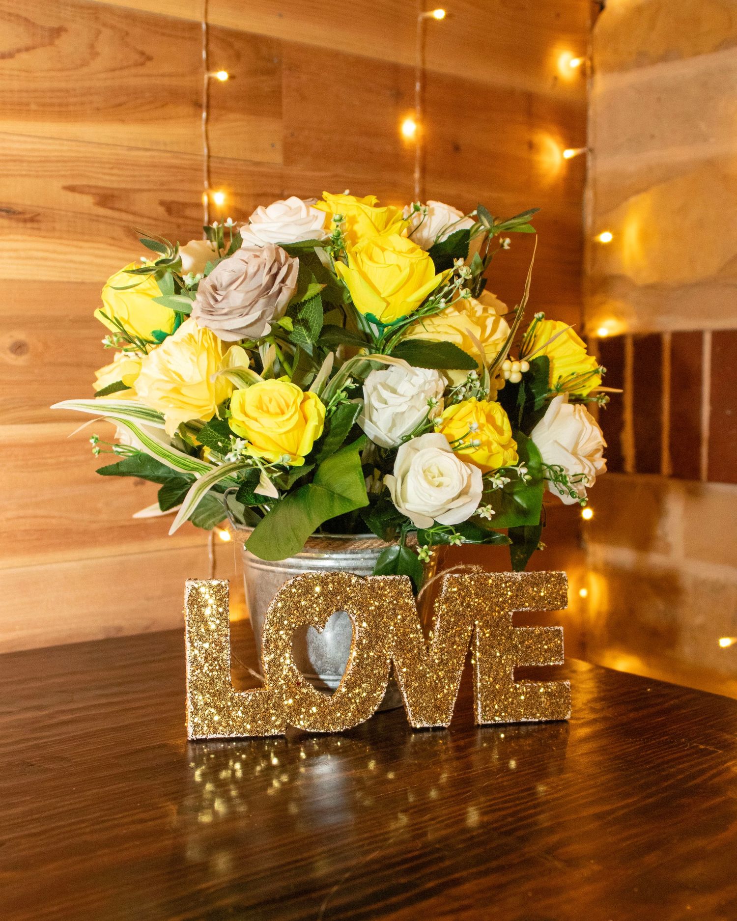 Yellow and white roses arranged in a vase with a gold glittery 'LOVE' sign decoration on a wooden table with twinkle lights.