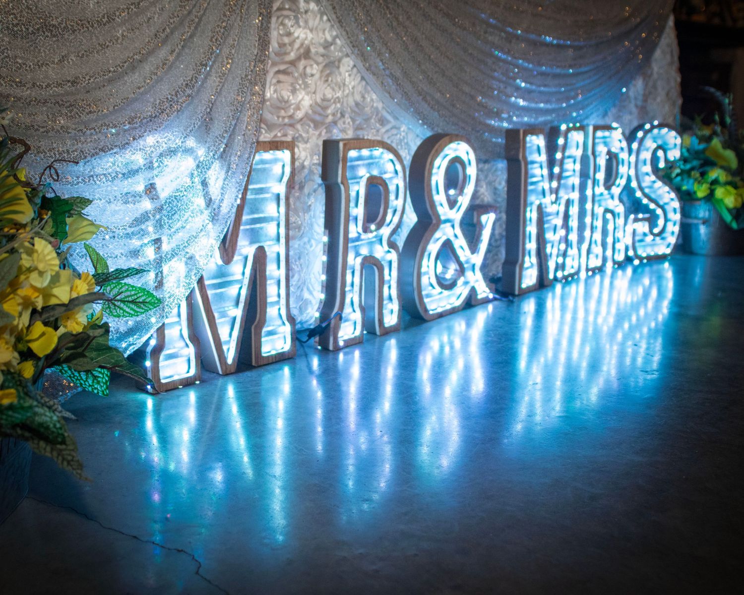 A glowing neon blue MR & MRS sign illuminates an elegant wedding venue backdrop with sheer white drapery.