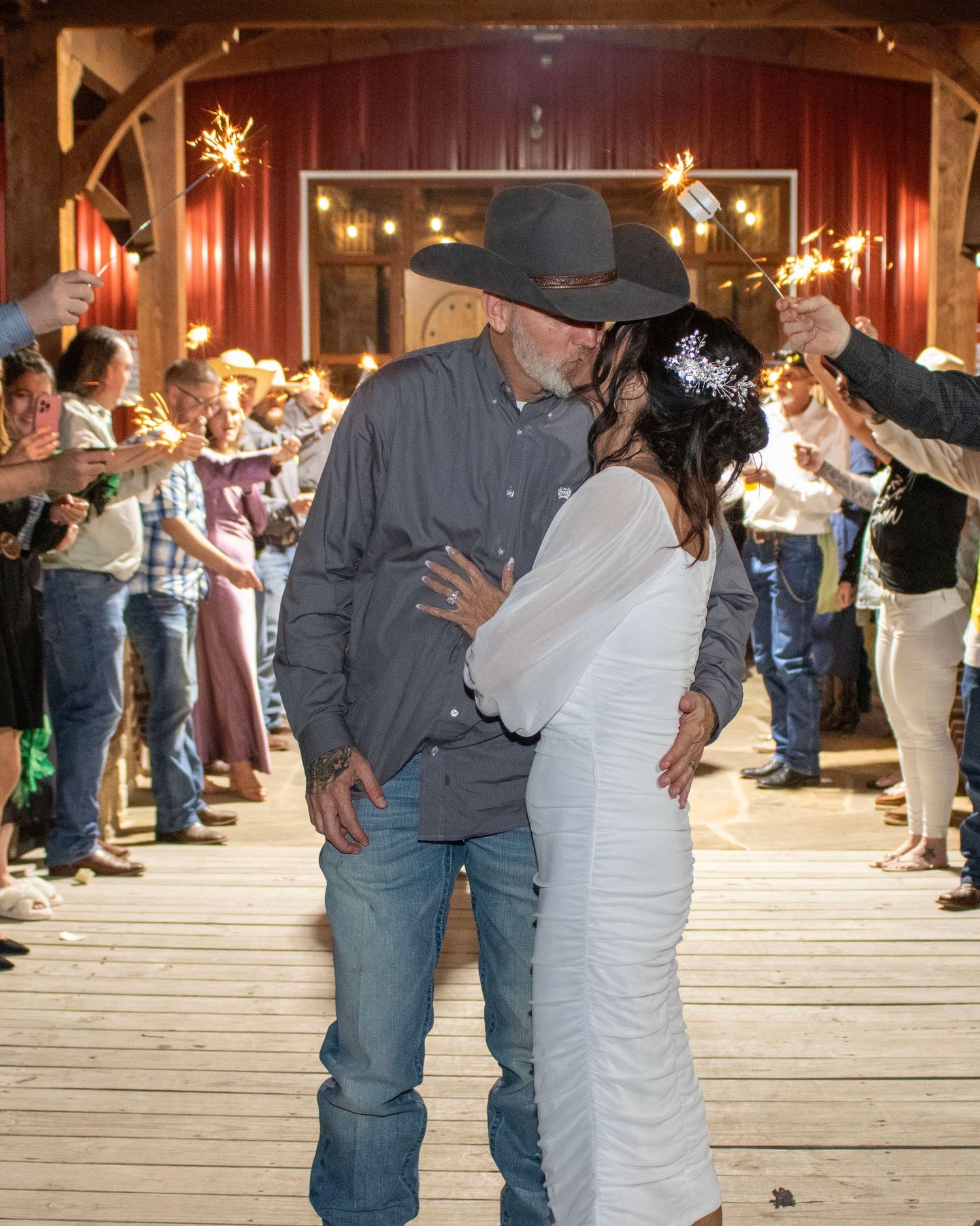 Cowboy-themed wedding couple embraces during a sparkler exit send-off in a rustic barn setting.
