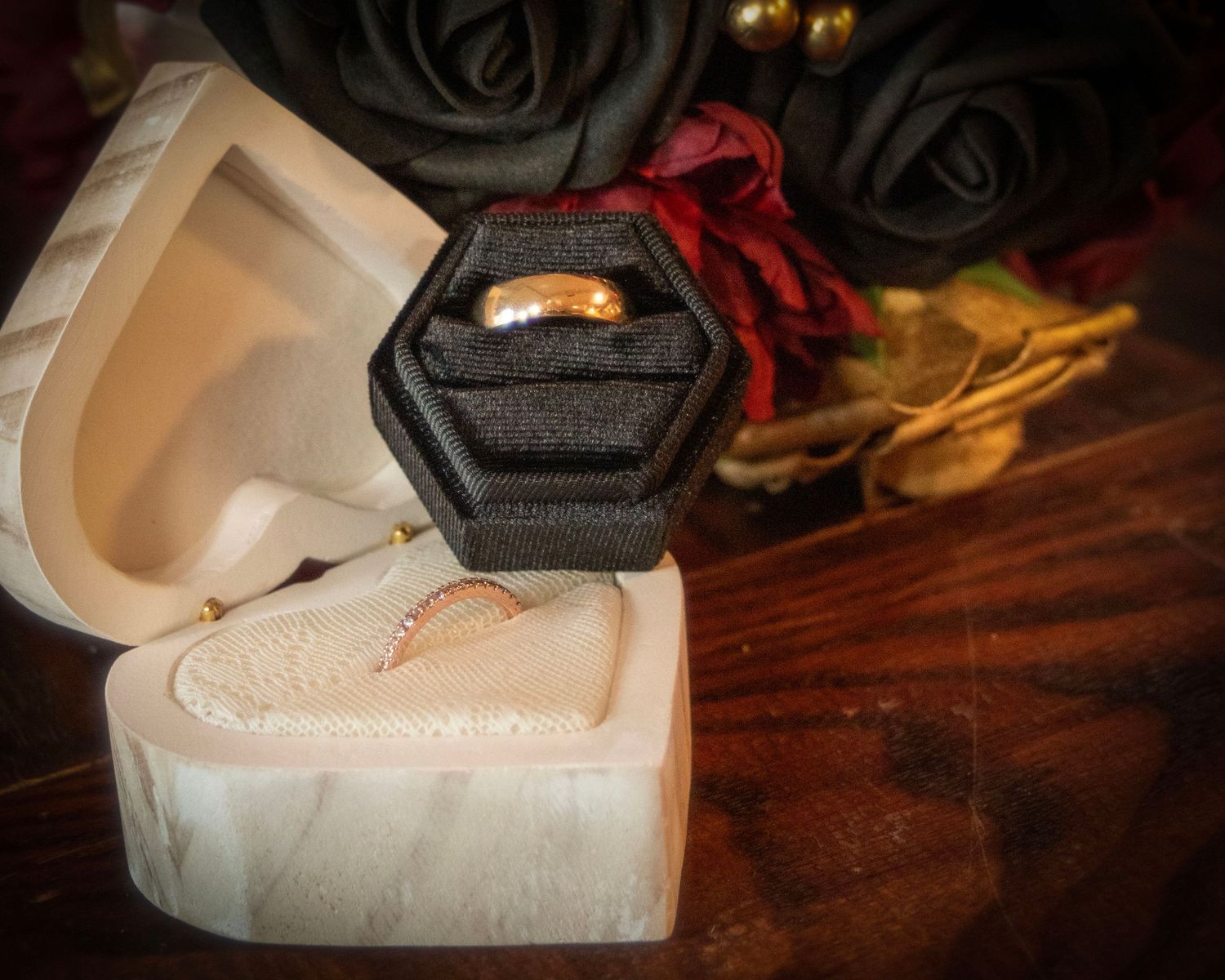 Gold wedding ring in black hexagonal box with burgundy flowers on dark wooden surface.