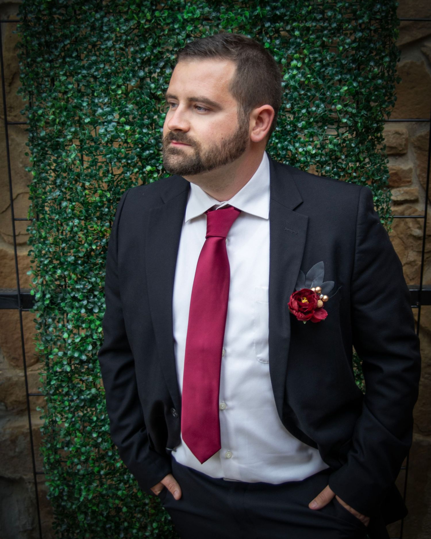 Well-dressed person in black suit and red tie poses against ivy-covered wall backdrop.