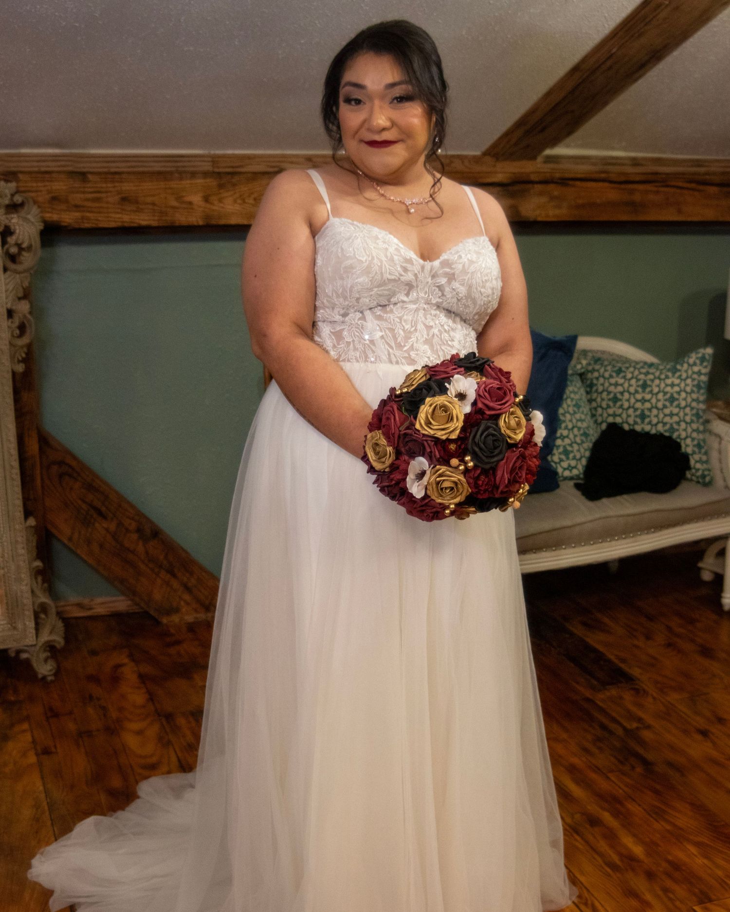 A bride in a white lace wedding gown holds a deep red and burgundy bouquet while standing in a rustic room with wooden beams.