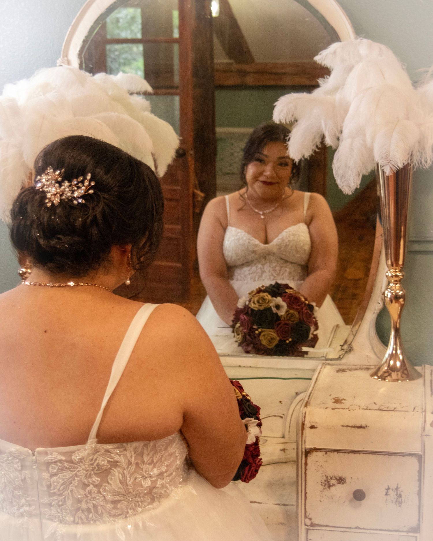 A bride wearing a white lace dress looks at herself in a vanity mirror adorned with white feather decorations.