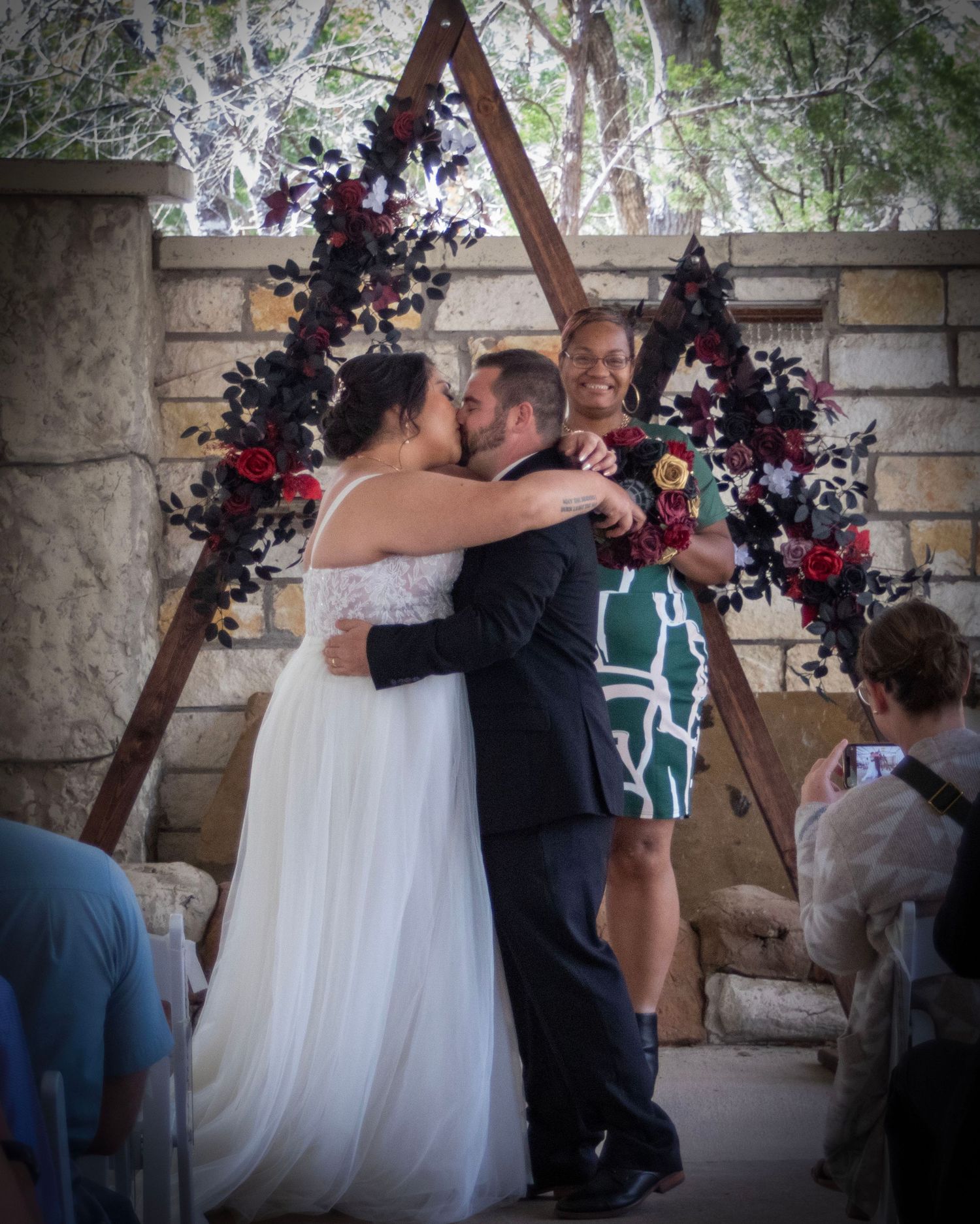 Newlyweds share their first kiss under a wooden triangle arch decorated with dark purple and red flowers.