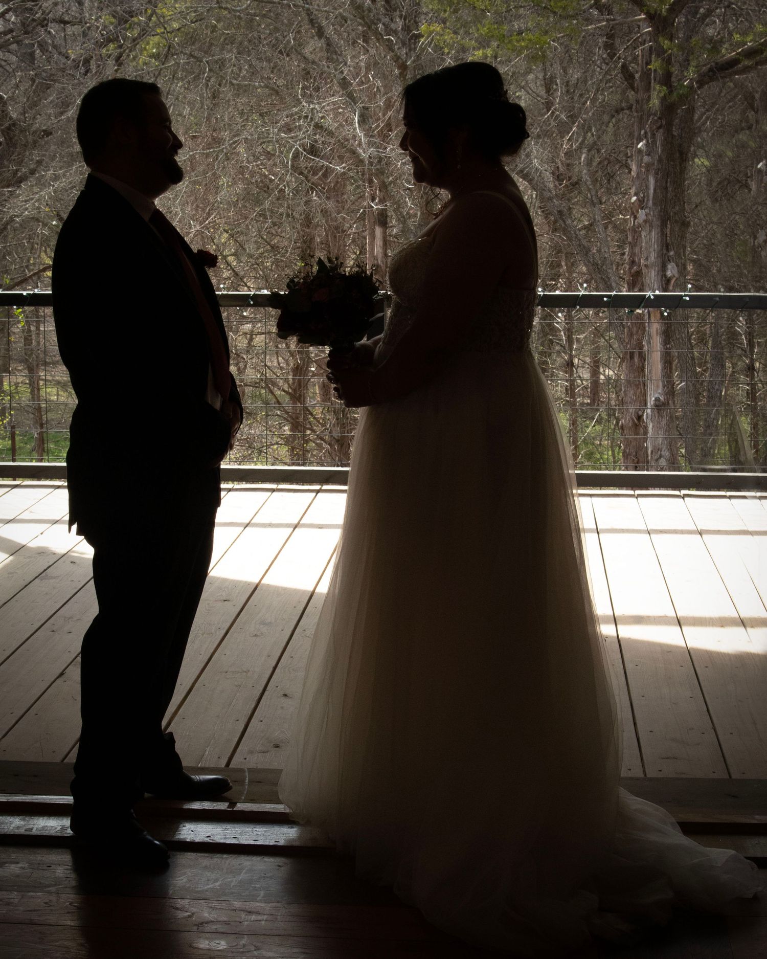 Silhouette of bride and groom standing together on a porch with sunlit trees in background.