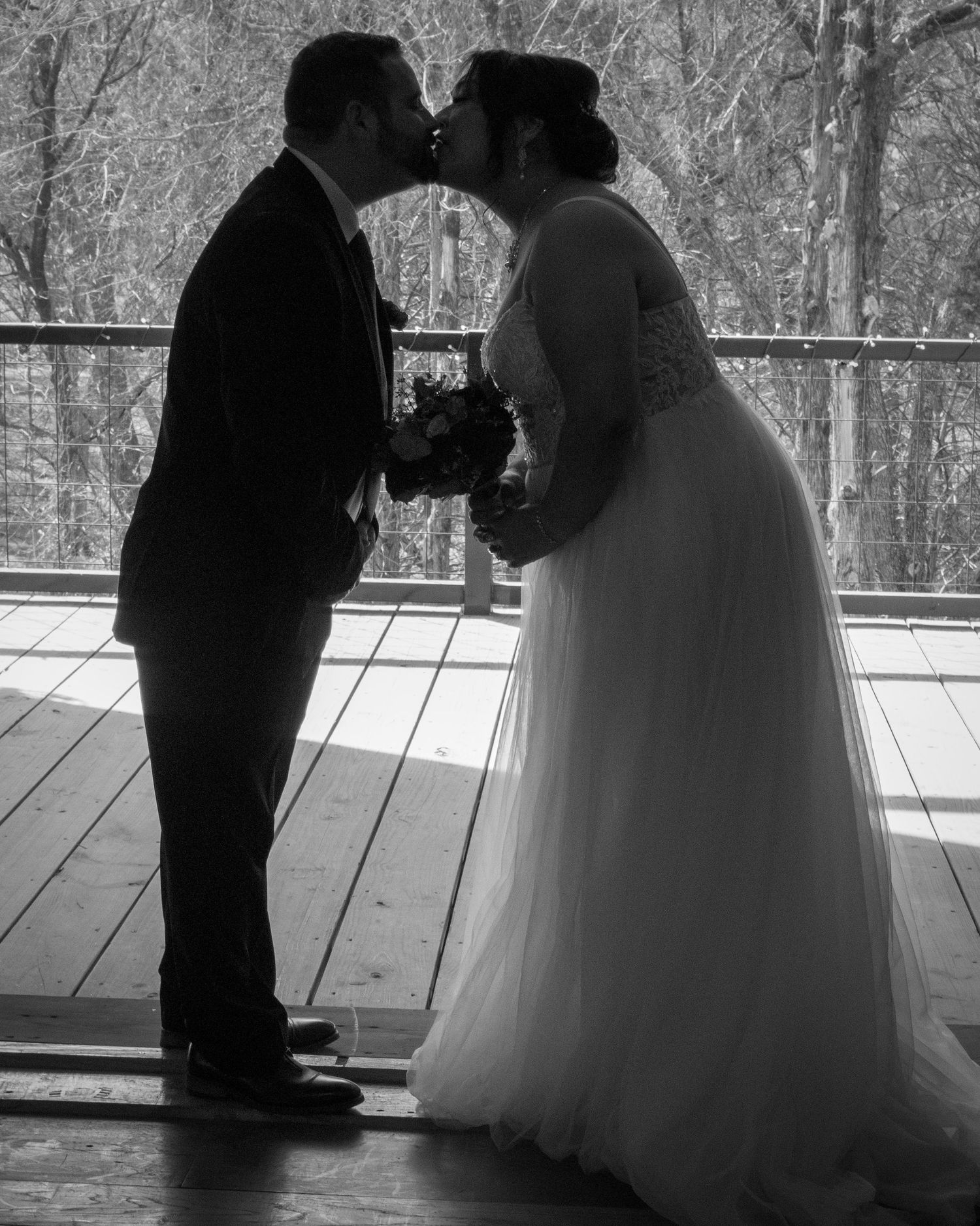 Black and white silhouette of newlyweds sharing romantic kiss on wooden deck surrounded by trees.