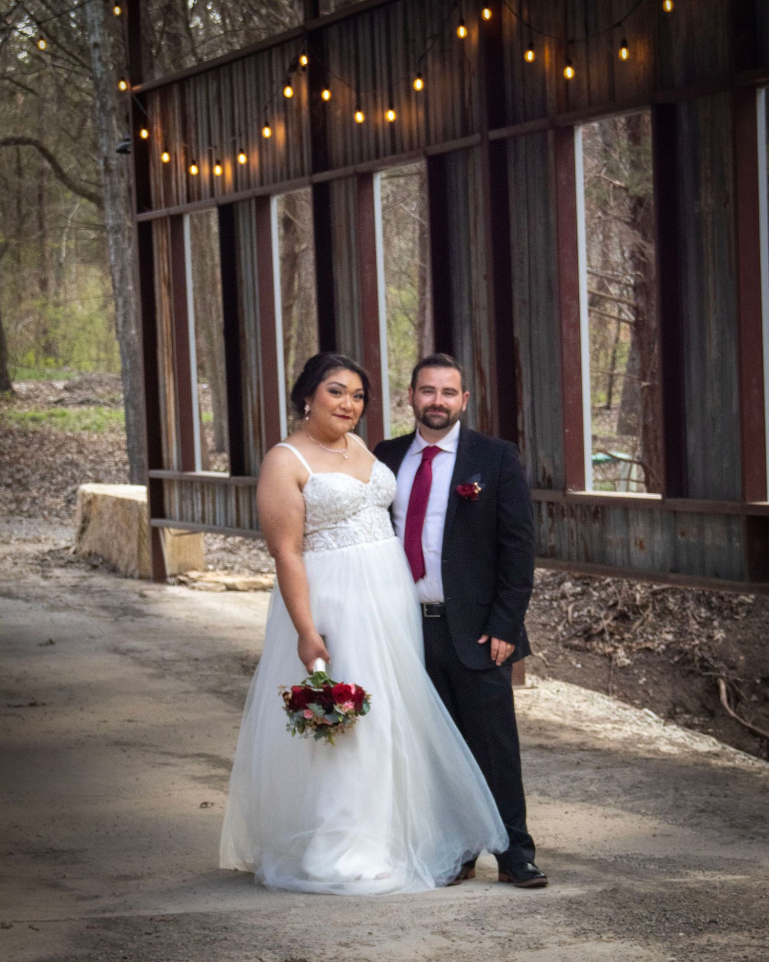 A couple poses under string lights in front of a rustic wooden barn on their wedding day.