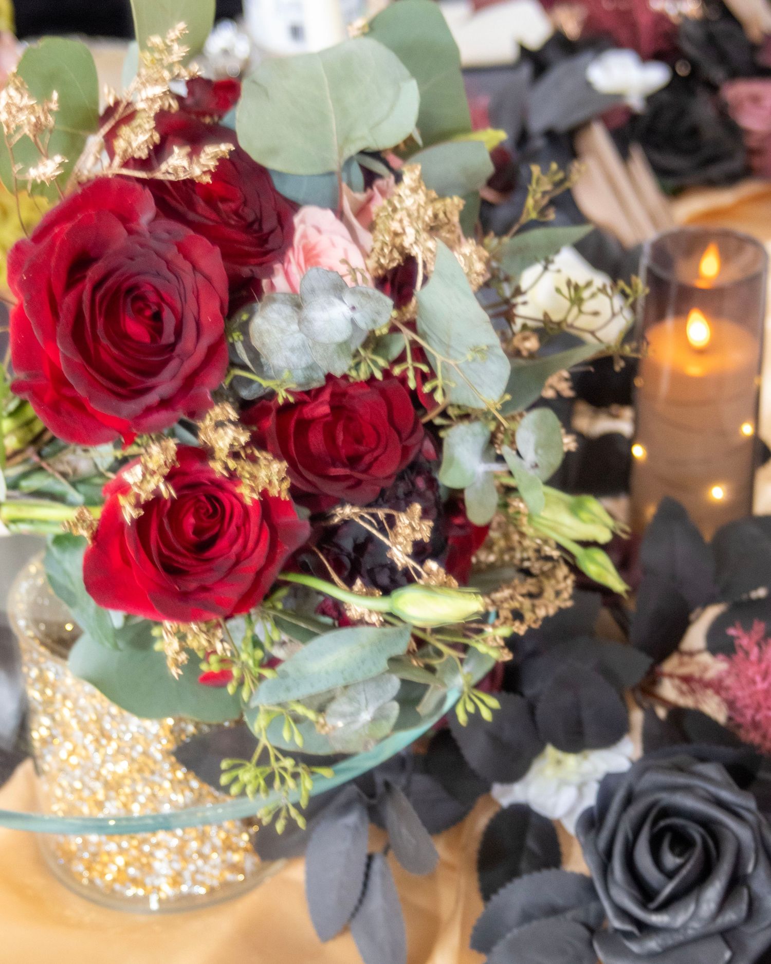 Elegant floral centerpiece with deep red roses and eucalyptus in a metallic gold vase next to a glowing candle.