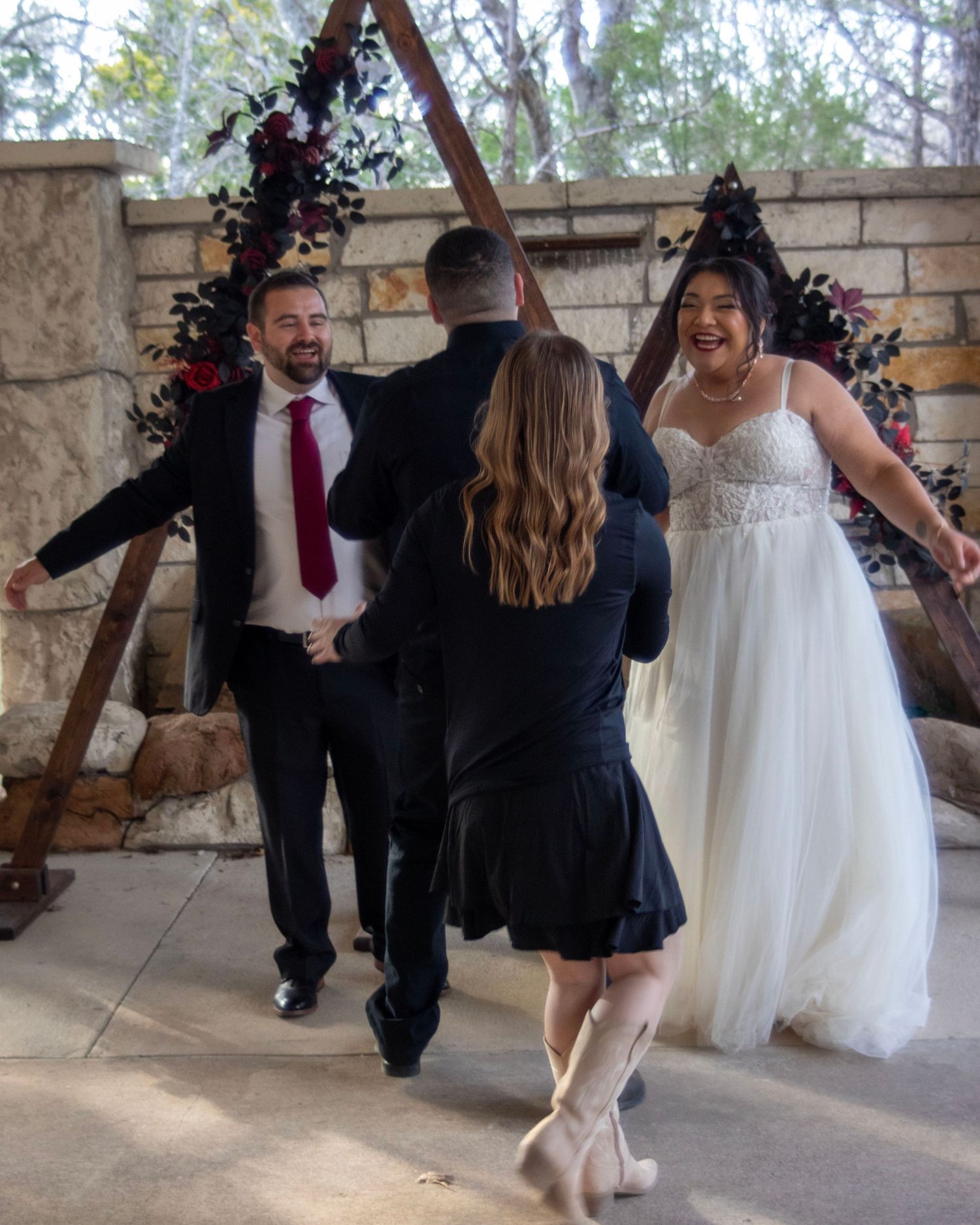 Wedding couple dances with guests under wooden arch decorated with flowers at outdoor stone venue.