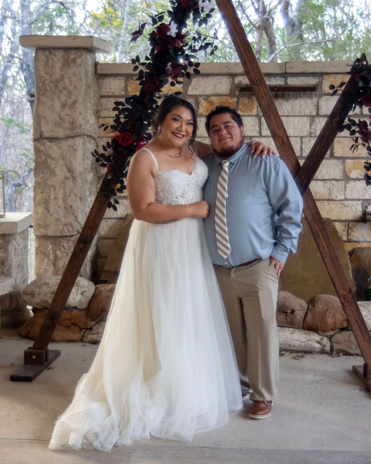 Outdoor wedding ceremony setup with wooden triangular arch decorated with dark flowers against stone wall backdrop.