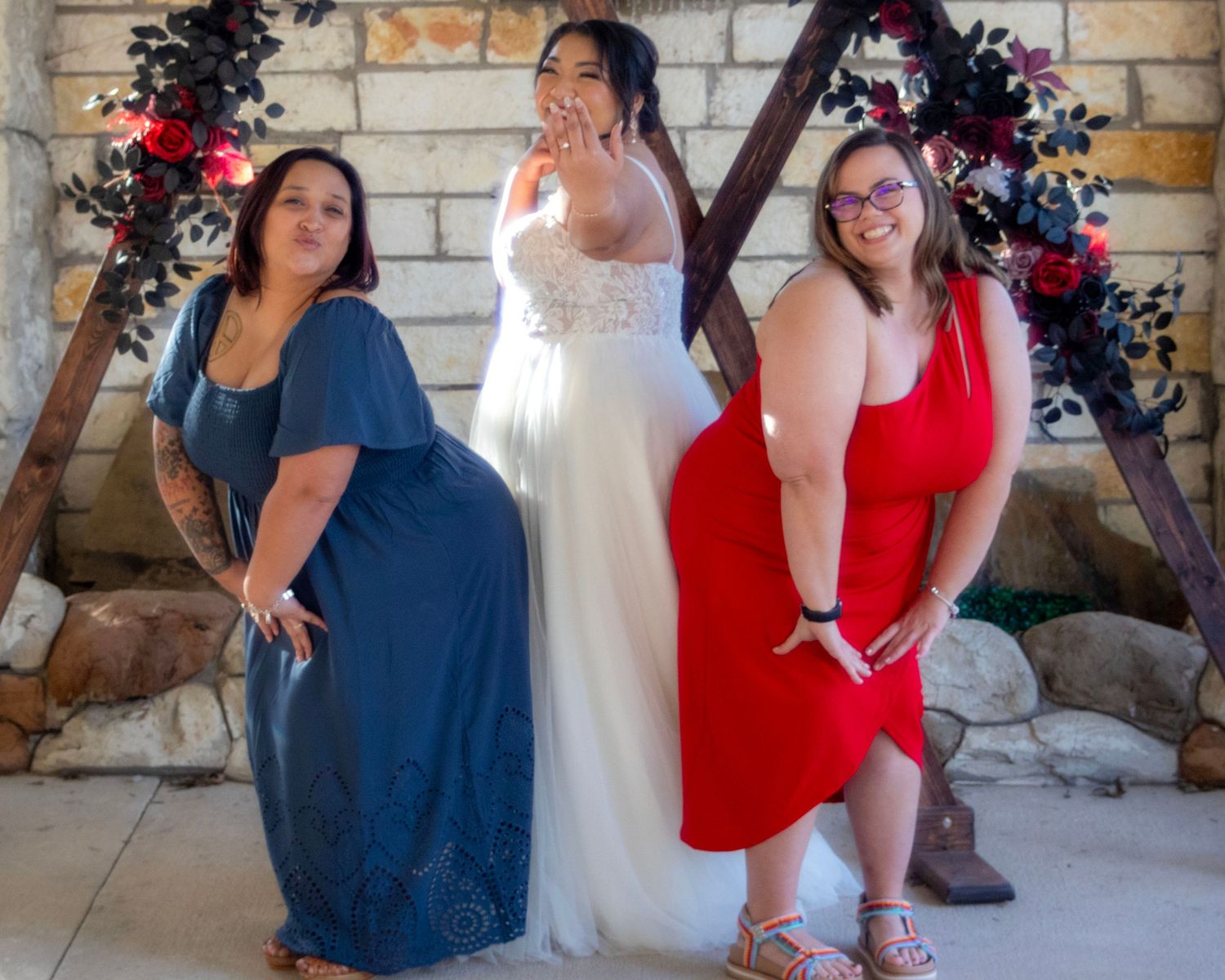 Three women in coordinating red, white and blue dresses pose together by a stone wall with floral decorations.