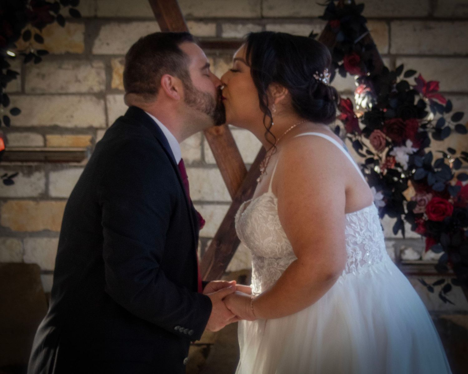 A tender moment between newlyweds against a stone wall adorned with red and pink flowers.