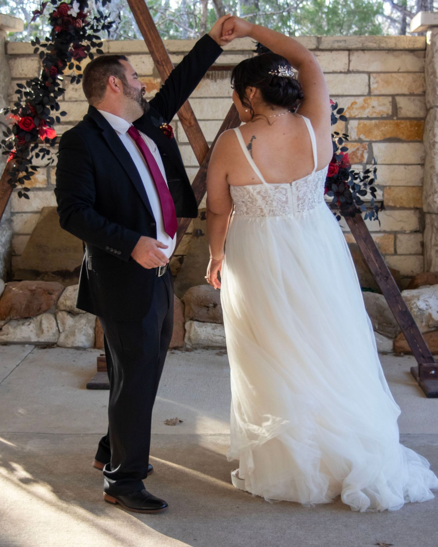 Dancing couple in wedding attire under a stone archway with red floral decor in outdoor setting.