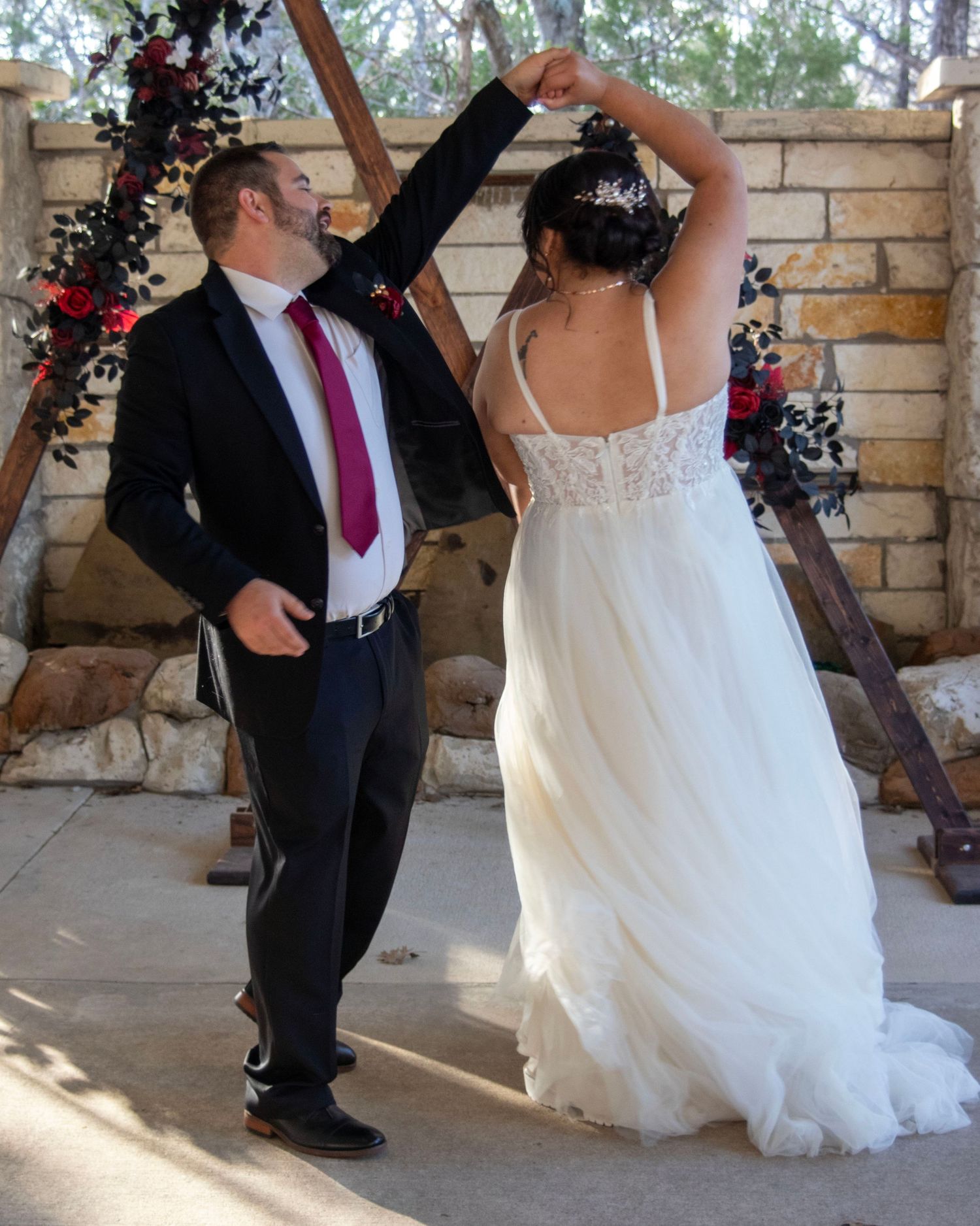 Dancing under a rustic wooden arch with stone wall backdrop during wedding celebration.