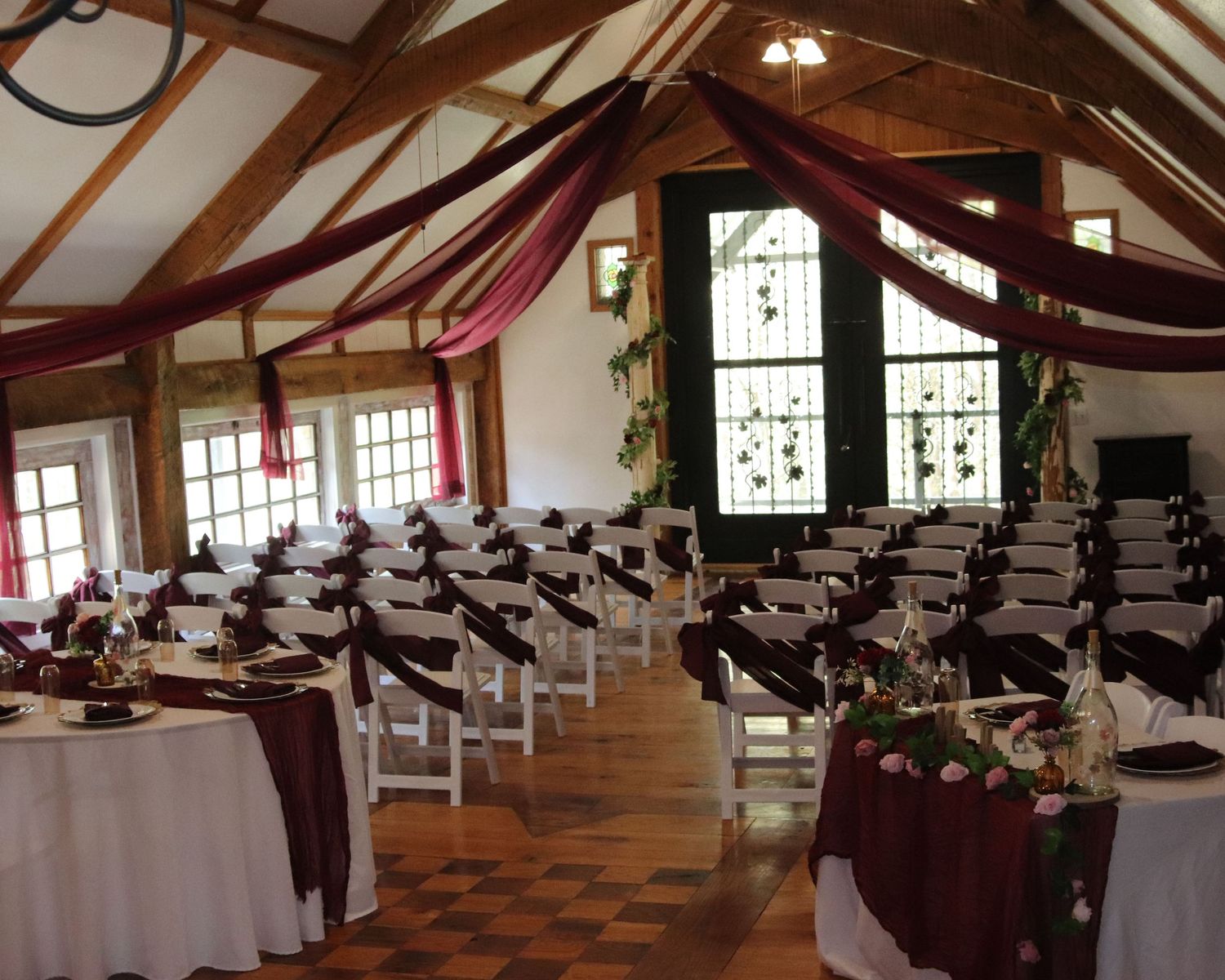 Indoor event venue with burgundy fabric draping, white chairs, and stained glass windows set up for a wedding ceremony.