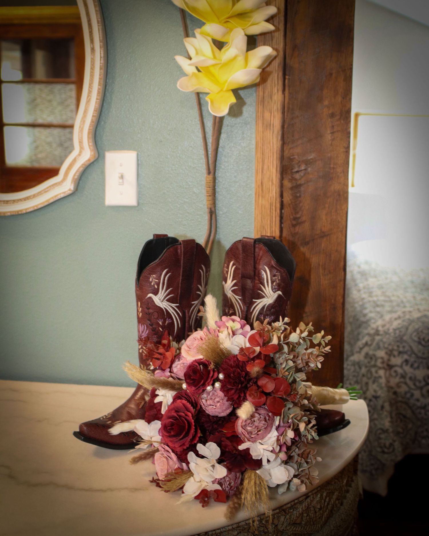 A pair of burgundy cowboy boots with white embroidery displayed with a pink and burgundy floral bouquet on a rustic table.