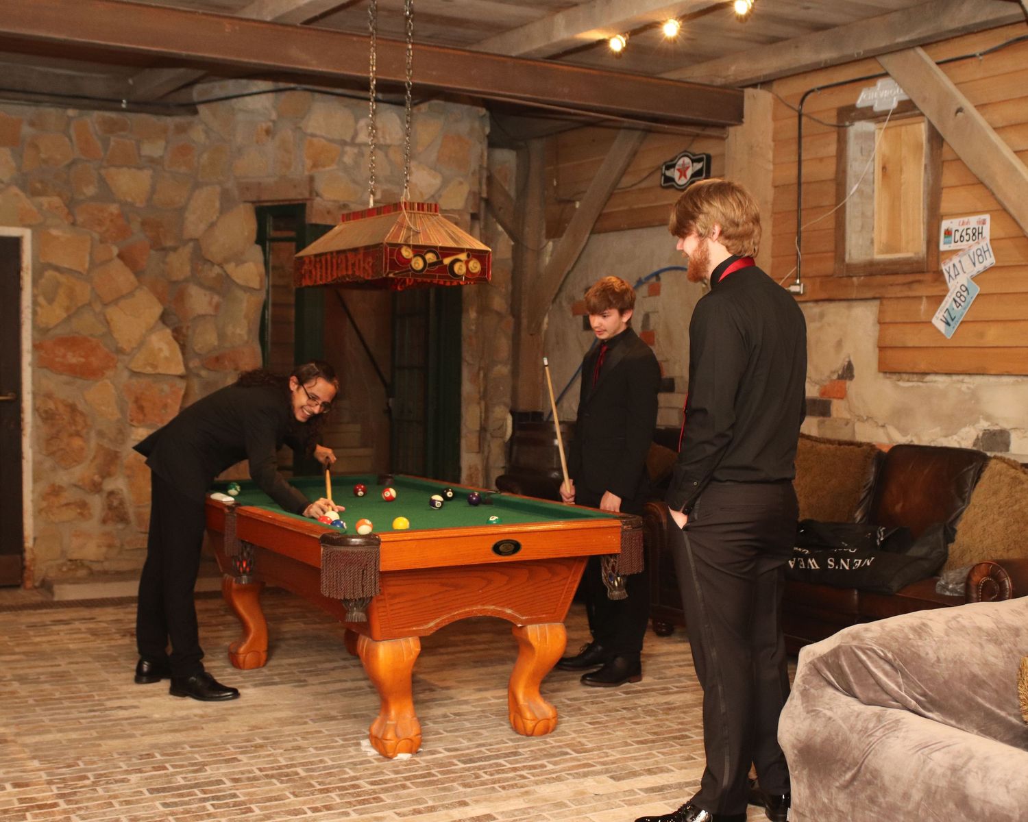 People playing a game of pool on an orange wooden table in a rustic basement room with stone walls.