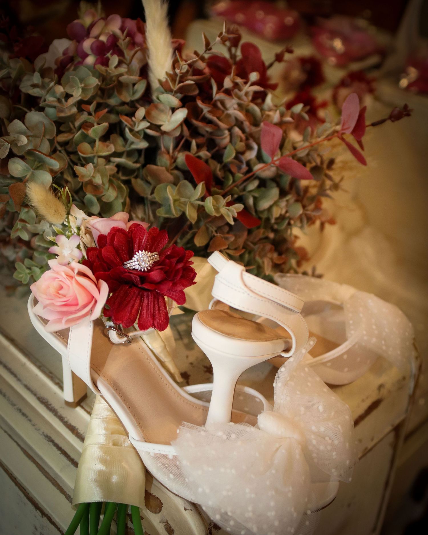 White bridal heels and a bouquet with red and pink flowers and dried eucalyptus arranged on a white surface.