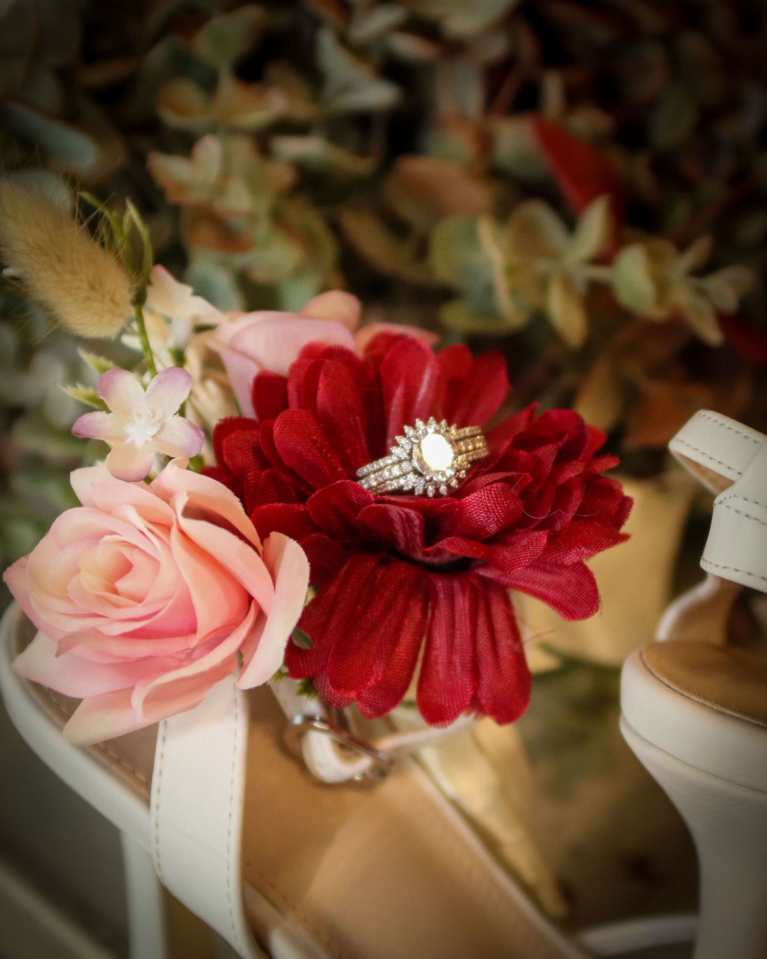 A diamond engagement ring nestles in a red dahlia flower next to a pink rose and white ribbon against a soft background.