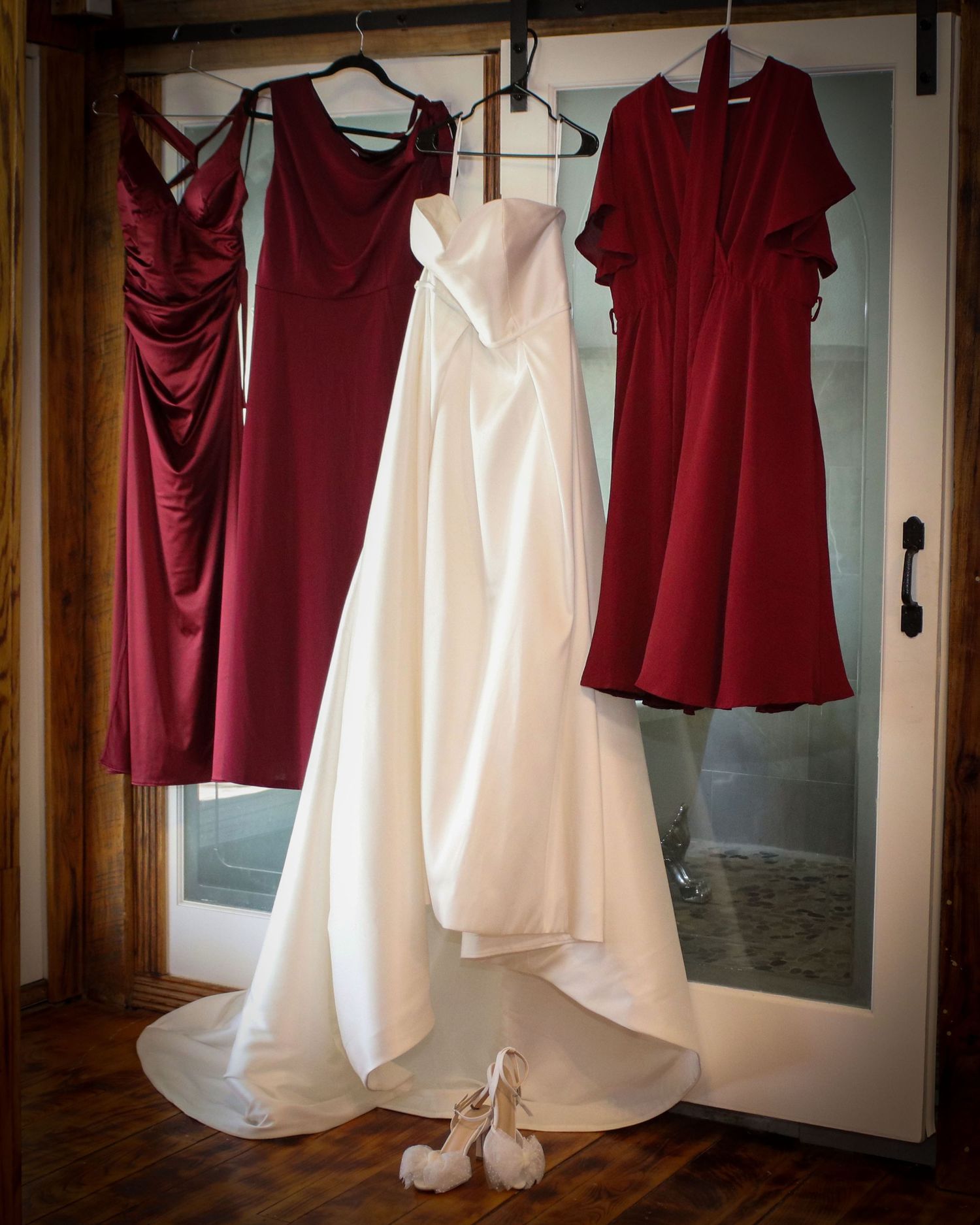 A white wedding dress hangs alongside three burgundy bridesmaid dresses with white bridal shoes on the floor.
