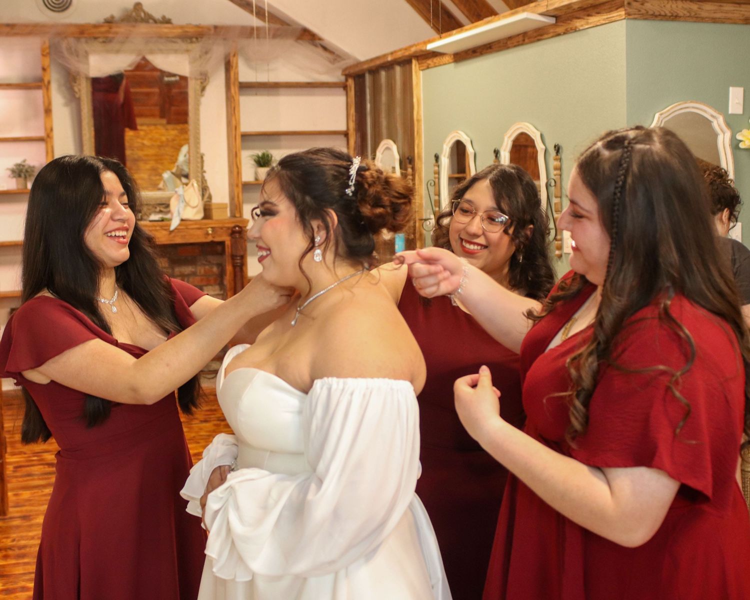 Bridesmaids in burgundy dresses help adjust the bride's strapless white gown in a rustic wooden venue.