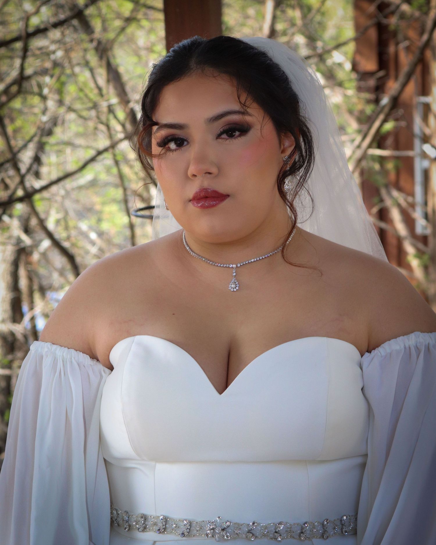 A bride in a classic white off-shoulder wedding dress with delicate bell sleeves and a crystal necklace poses outdoors.
