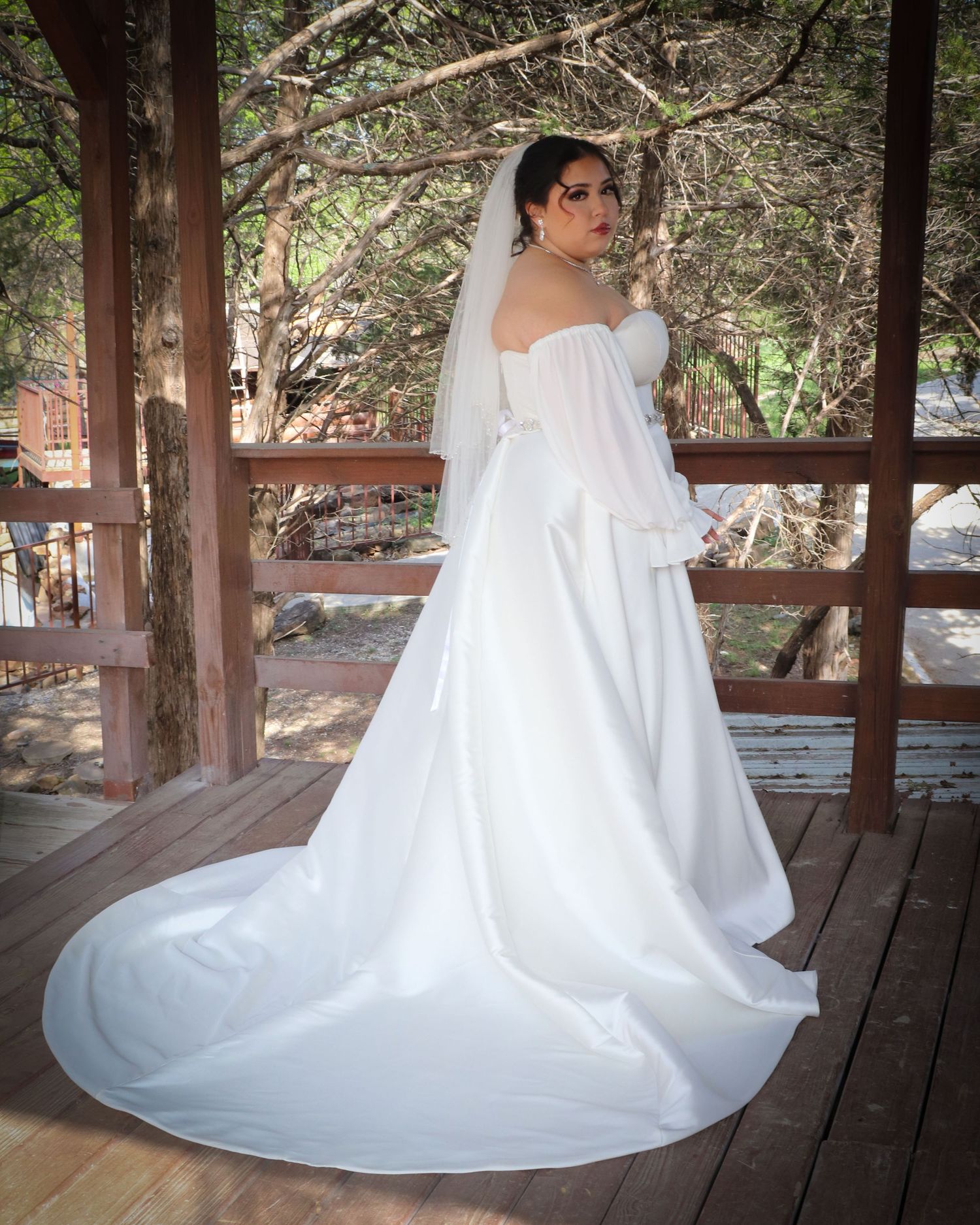 A bride in a flowing white wedding gown with long train and veil poses on a rustic wooden gazebo deck.
