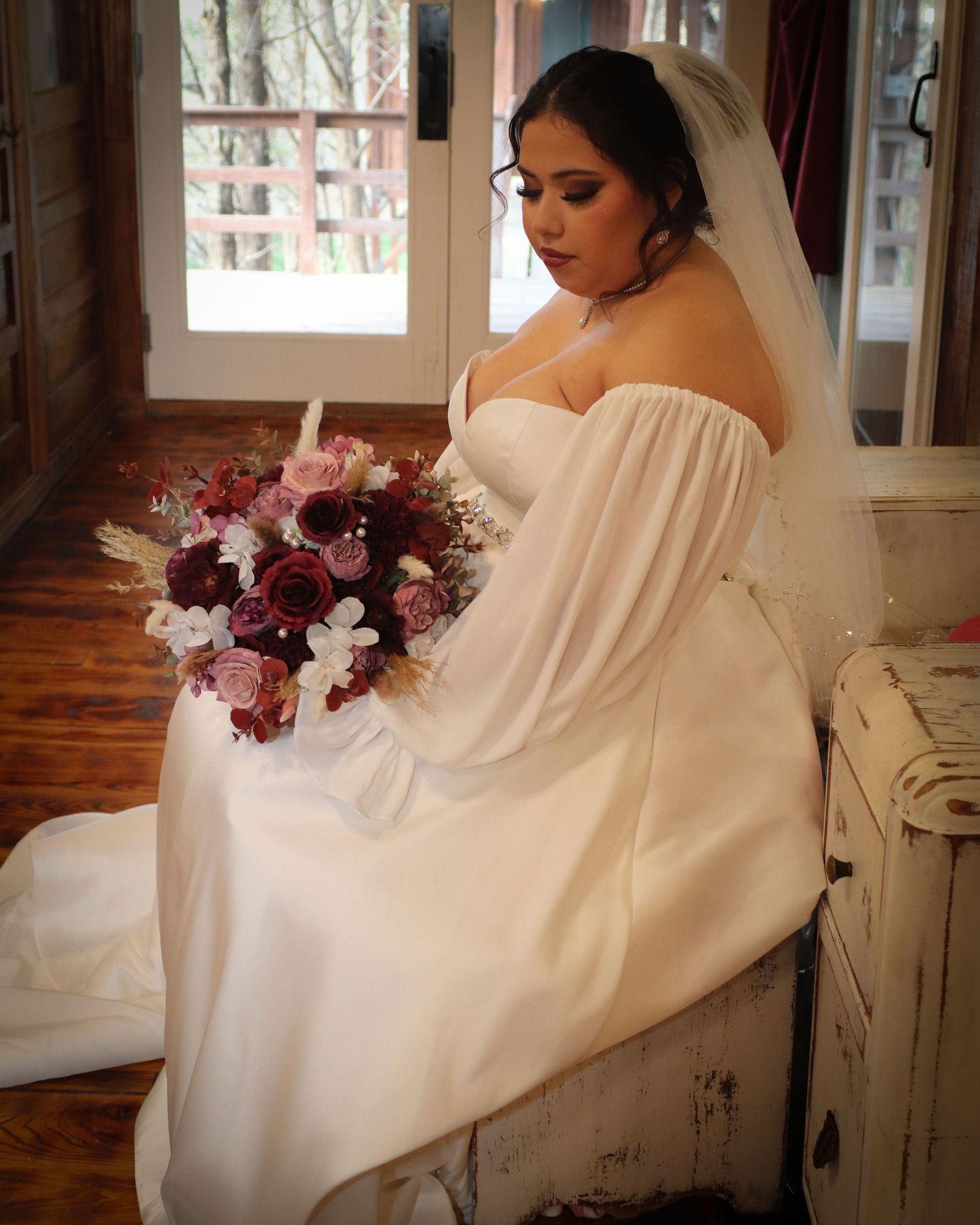 A bride in an elegant off-shoulder white dress holds a stunning burgundy and pink floral bouquet while seated near a window.