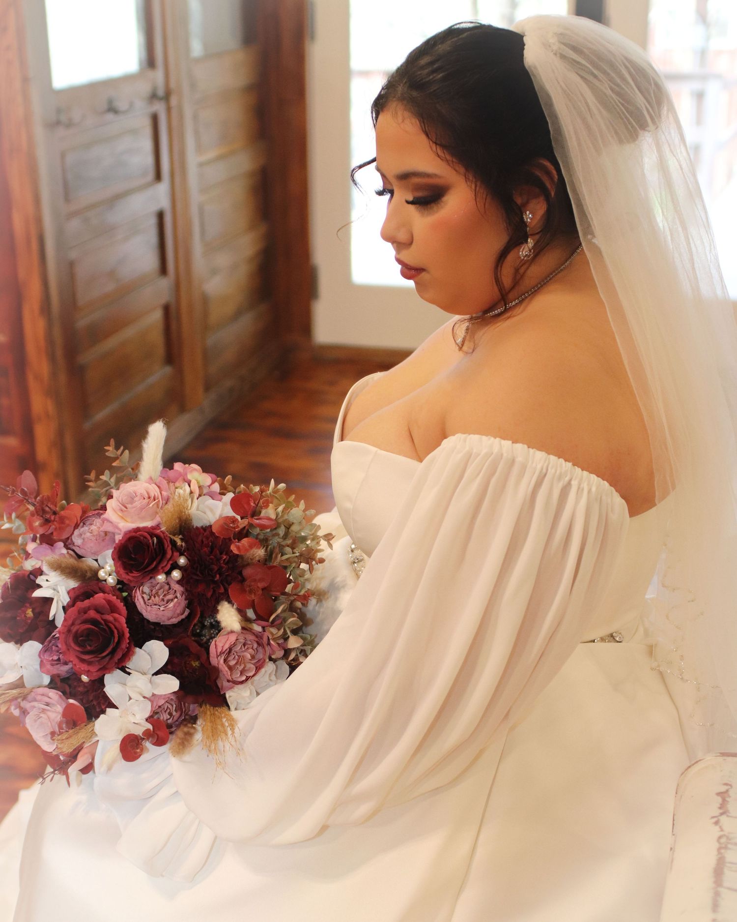 A bride in a flowing off-shoulder white dress poses with a stunning burgundy and pink floral bouquet.